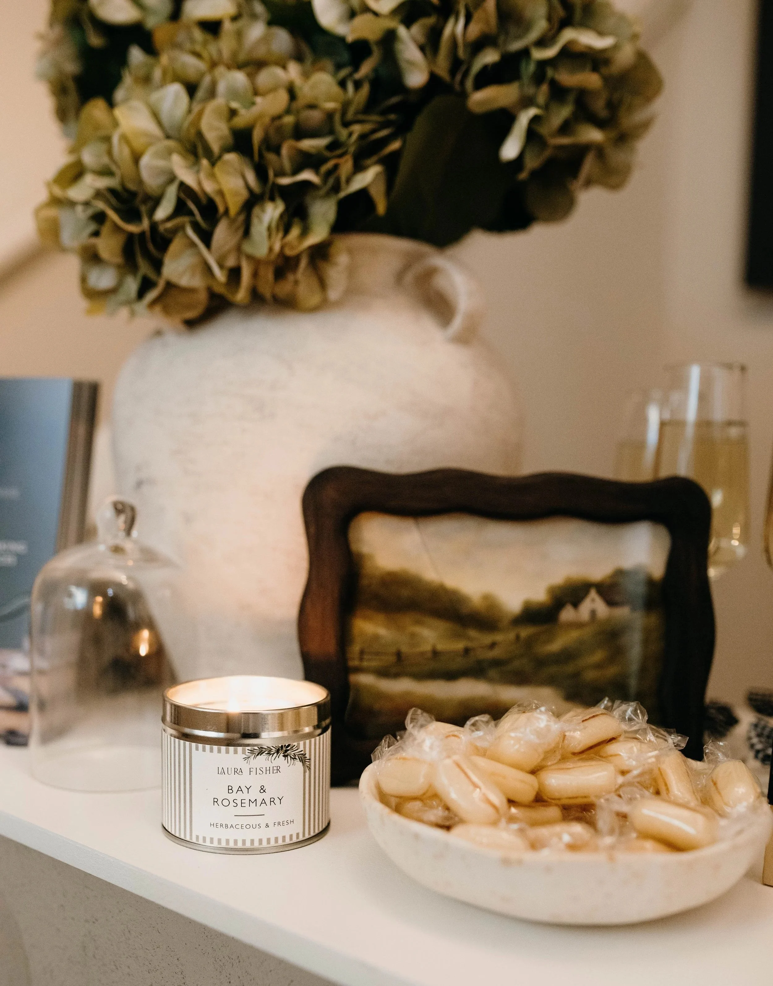 A white shelf with a scented candle labeled 'Laura Fisher Bay & Rosemary Herbaceous & Fresh,' a glass cloche covering a small object, and a plate of strips of soap or similar product in the foreground, a decorative vase with dried hydrangea flowers o