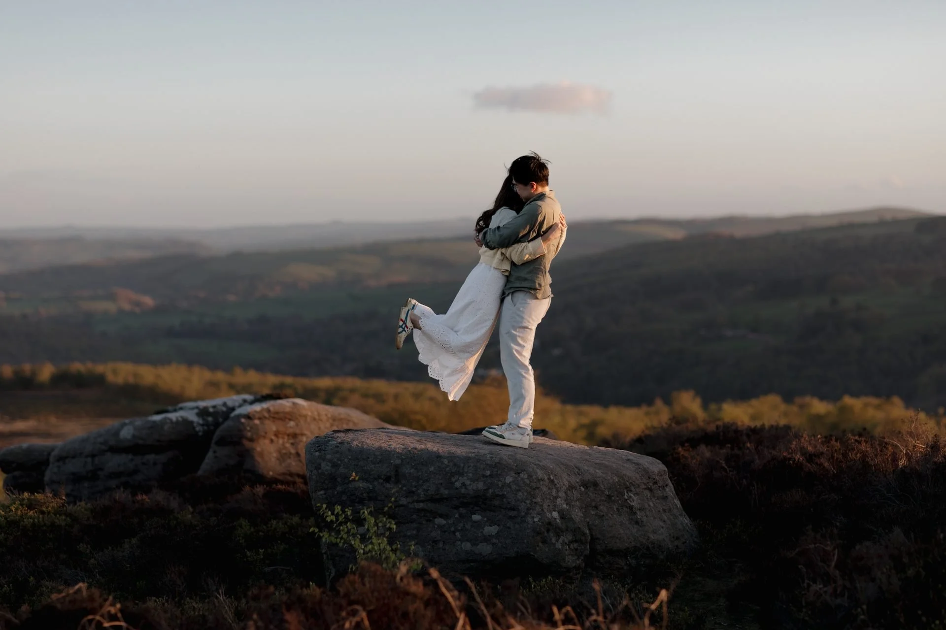 A couple embracing on a rocky hilltop with a scenic landscape of rolling hills and a cloudy sky in the background during sunset.