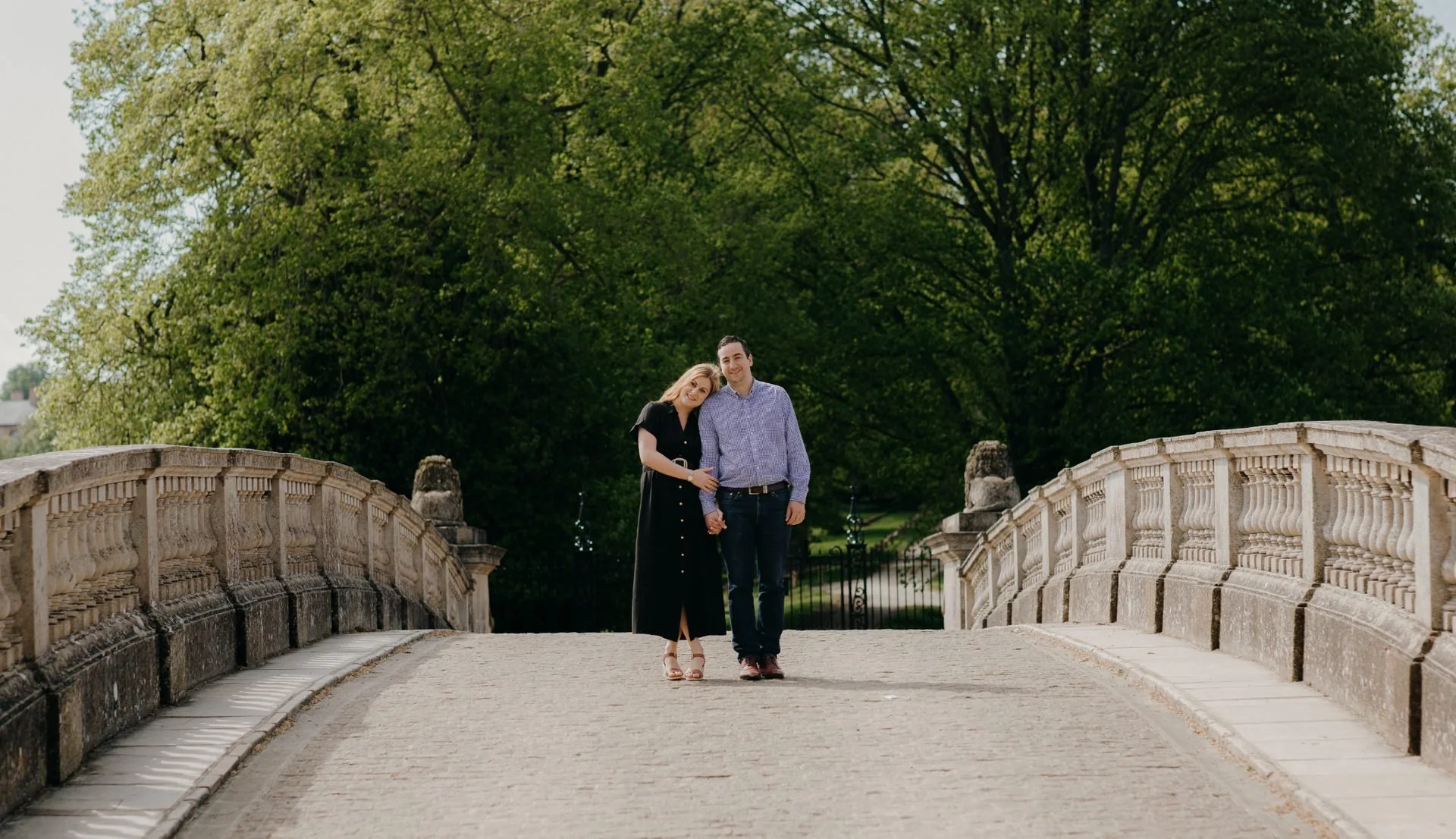 A couple walking across a stone bridge in a park, holding hands, with lush green trees in the background.