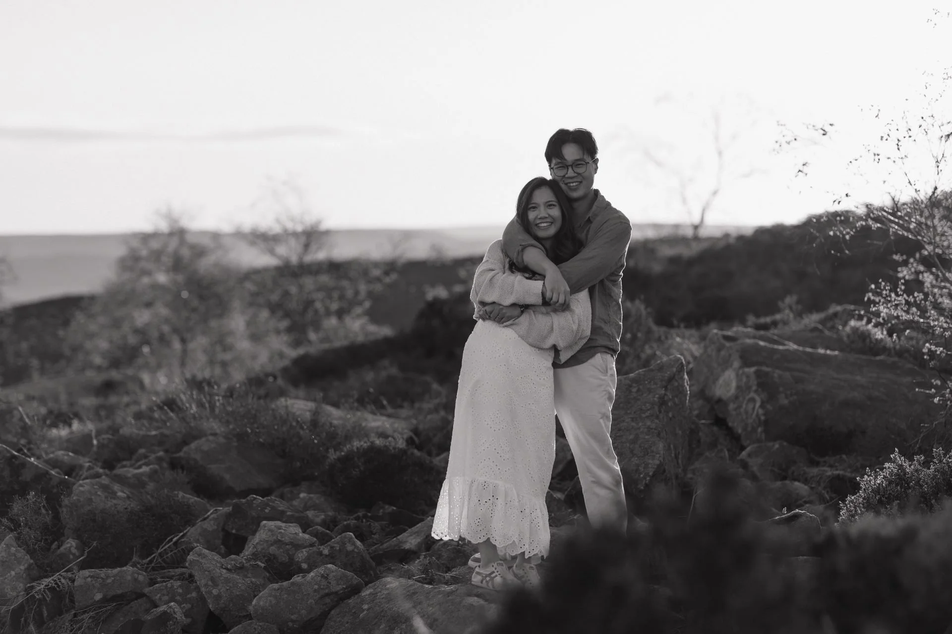 A black and white photo of a happy couple embracing outdoors. The woman is wearing a long, embroidered dress, and the man is in light-colored pants and a button-up shirt. They are smiling and standing on rocky terrain with trees and open land in the 