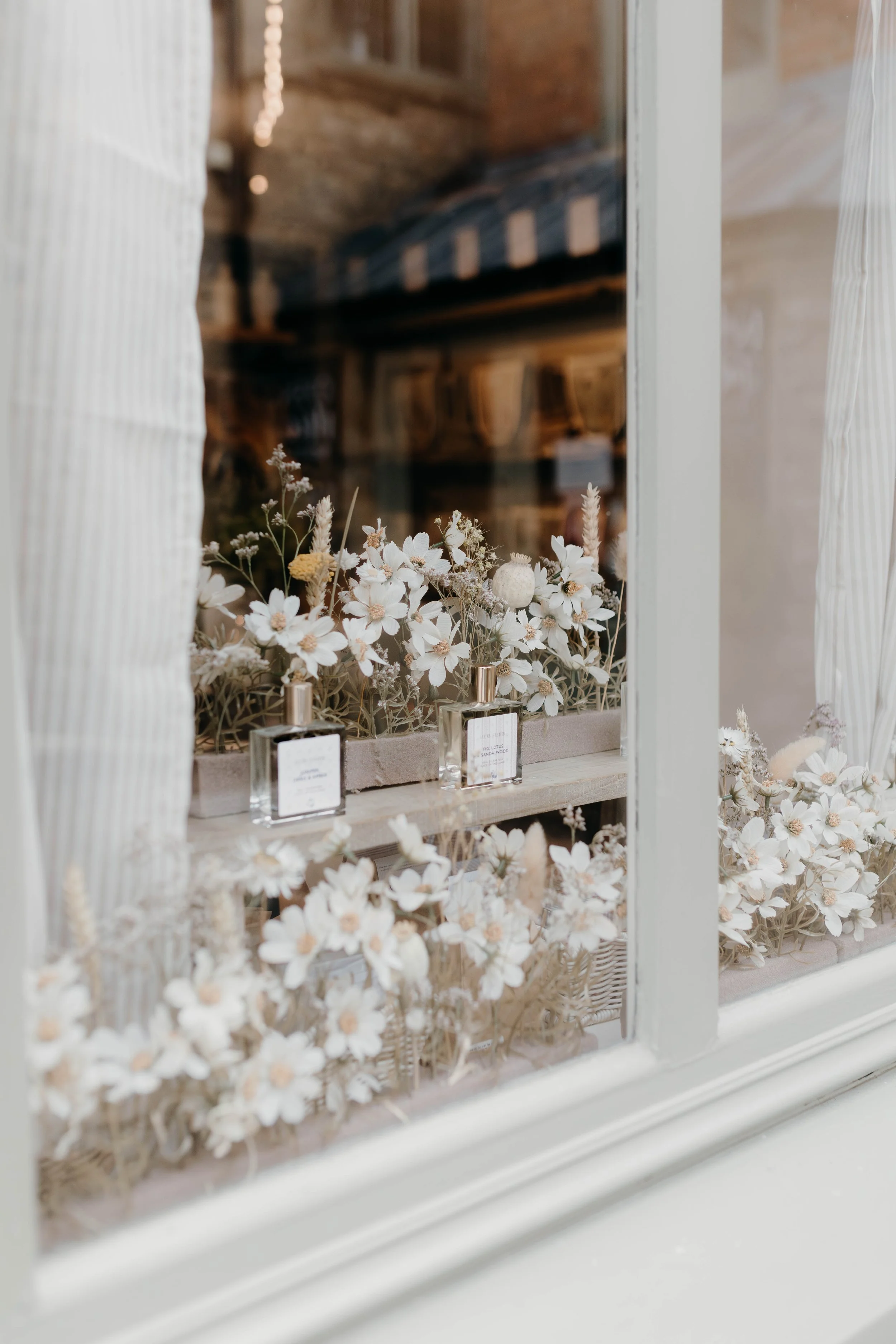 A window with white frame, decorated with white and beige flowers in a beige container, with small perfume bottles in front, inside a cozy room with exposed brick wall and string lights.
