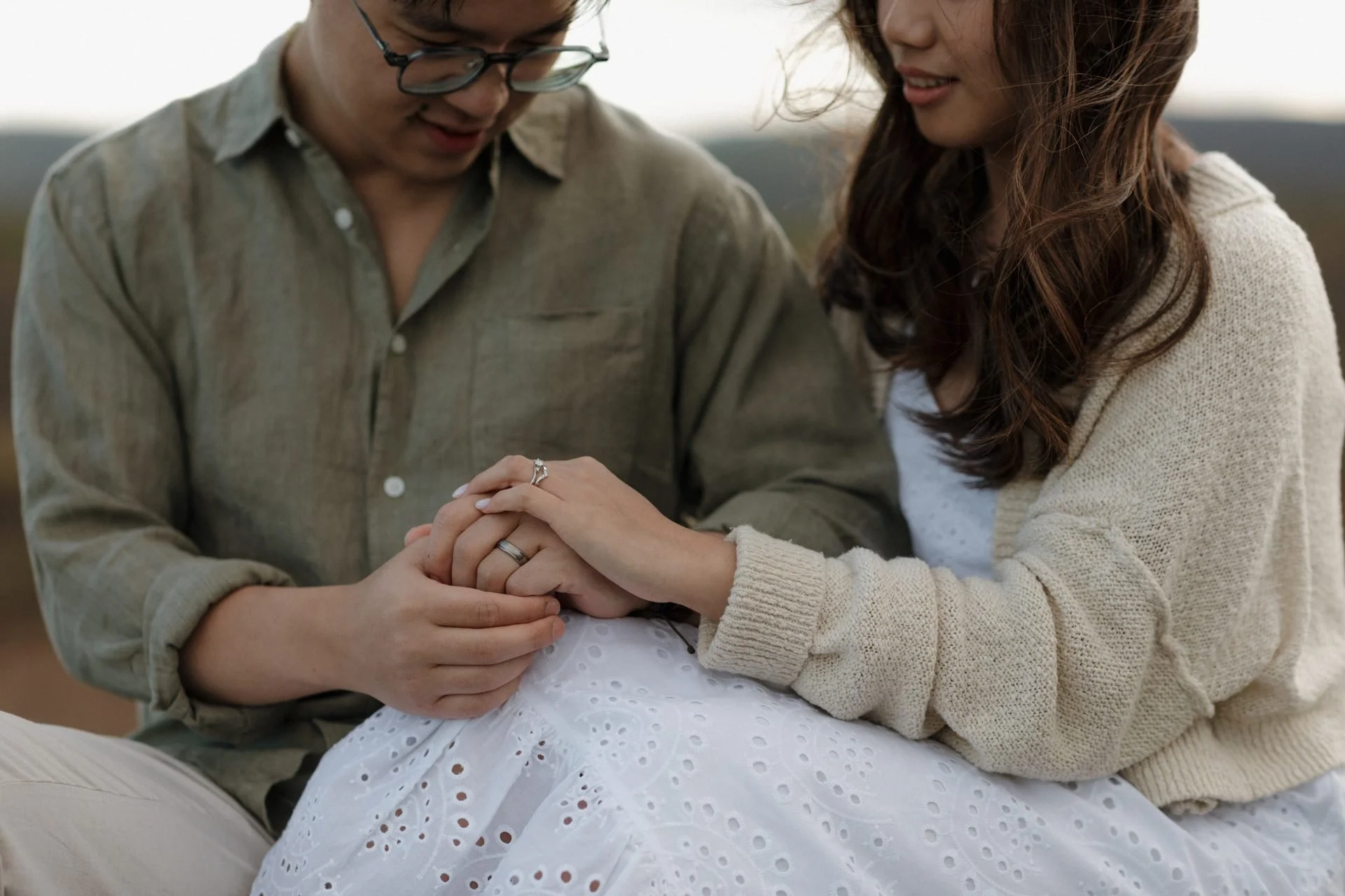 A couple holding hands during a baby shower or gender reveal event, with a pregnant woman in a white dress and a man in a green shirt, both smiling.
