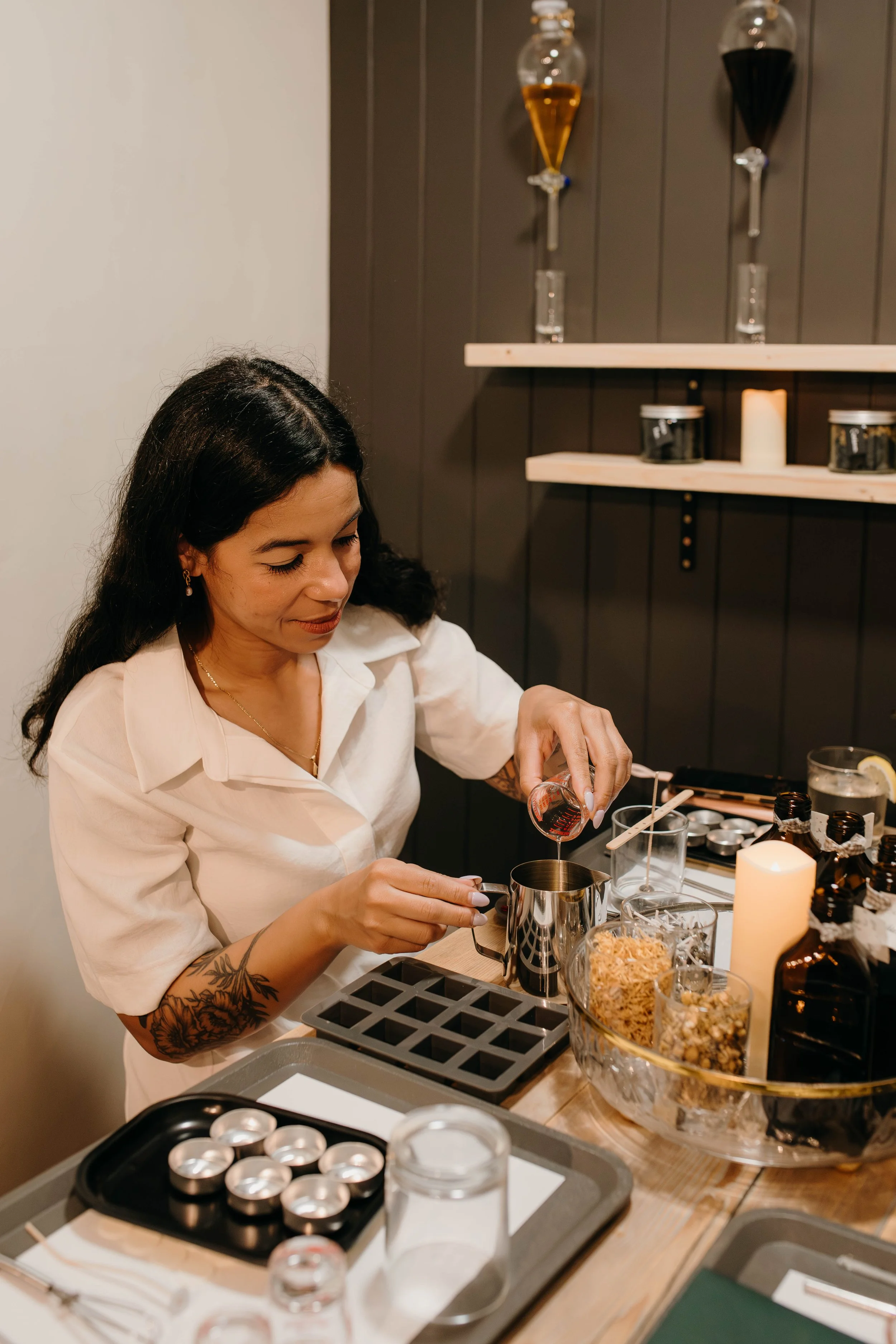 A woman with dark hair and tattoos on her arm is preparing ingredients in a kitchen, pouring liquid into a small metal pitcher surrounded by baking supplies, containers, and a candle on a wooden table.