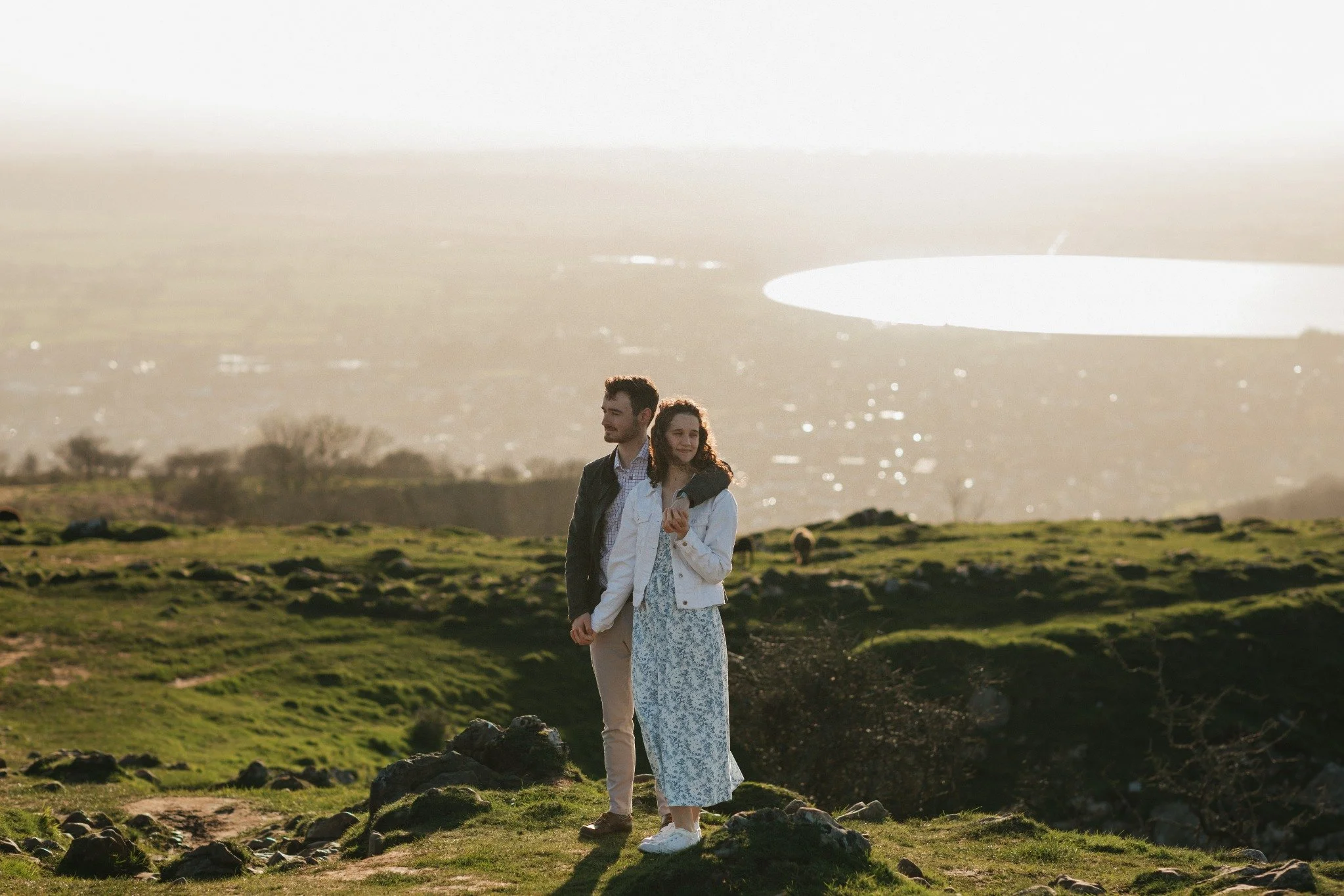 A couple walking outdoors on a grassy hillside during sunset, with a lake and distant hills in the background.