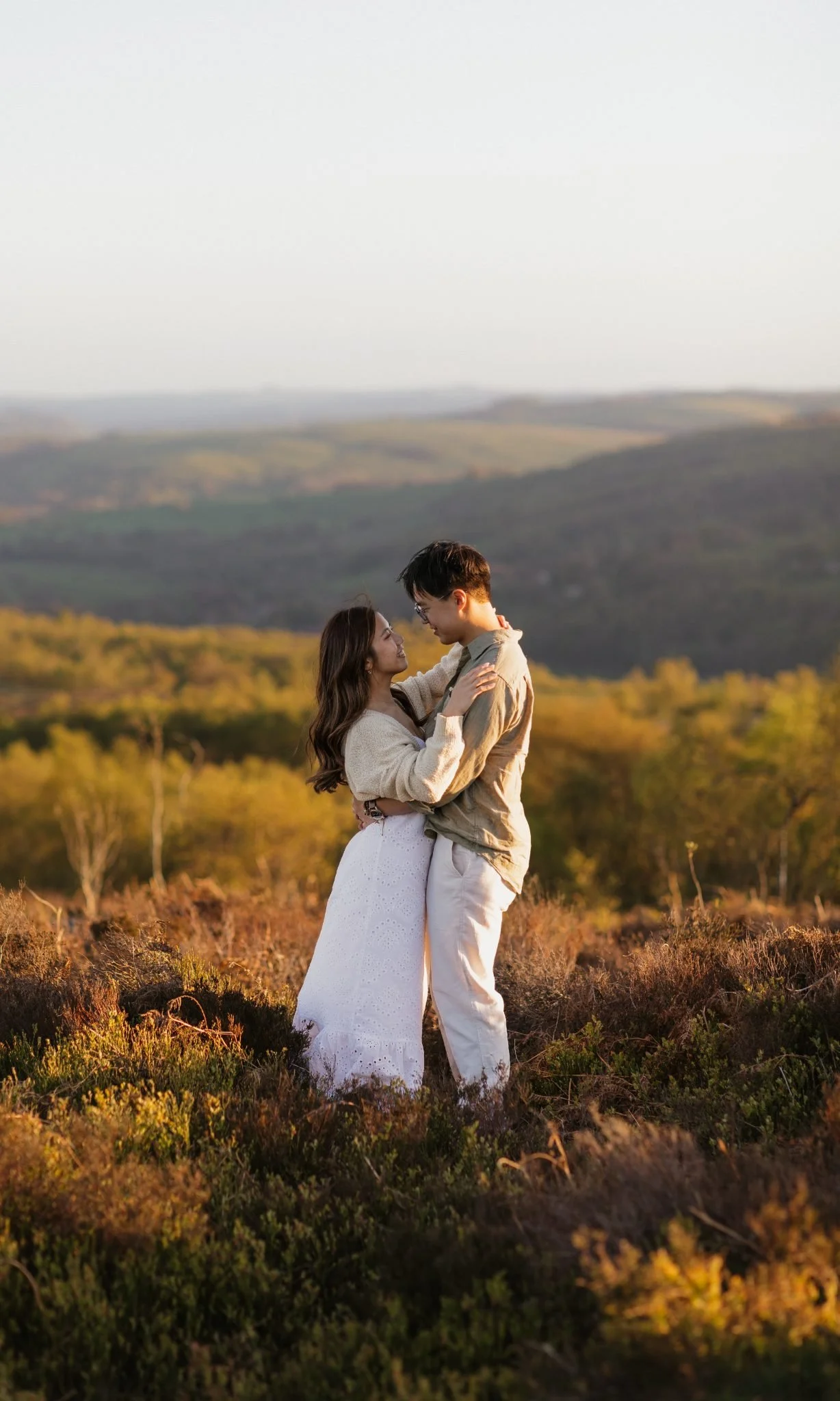 A couple stands close together outdoors in a scenic landscape with rolling hills and trees, embracing and smiling at each other during sunset.