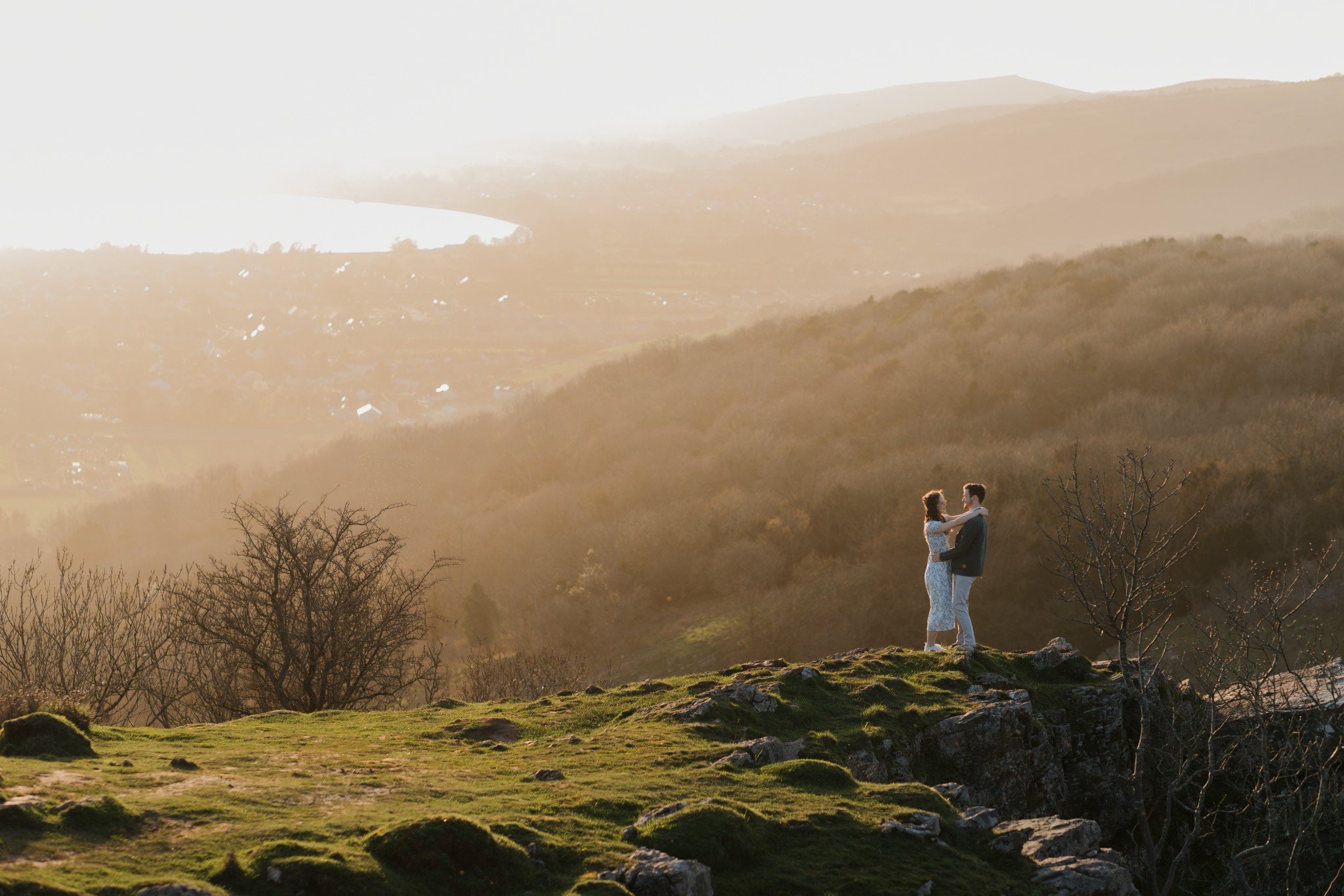 A couple standing and embracing on a grassy hilltop with a scenic mountain landscape in the background, bathed in warm sunlight.