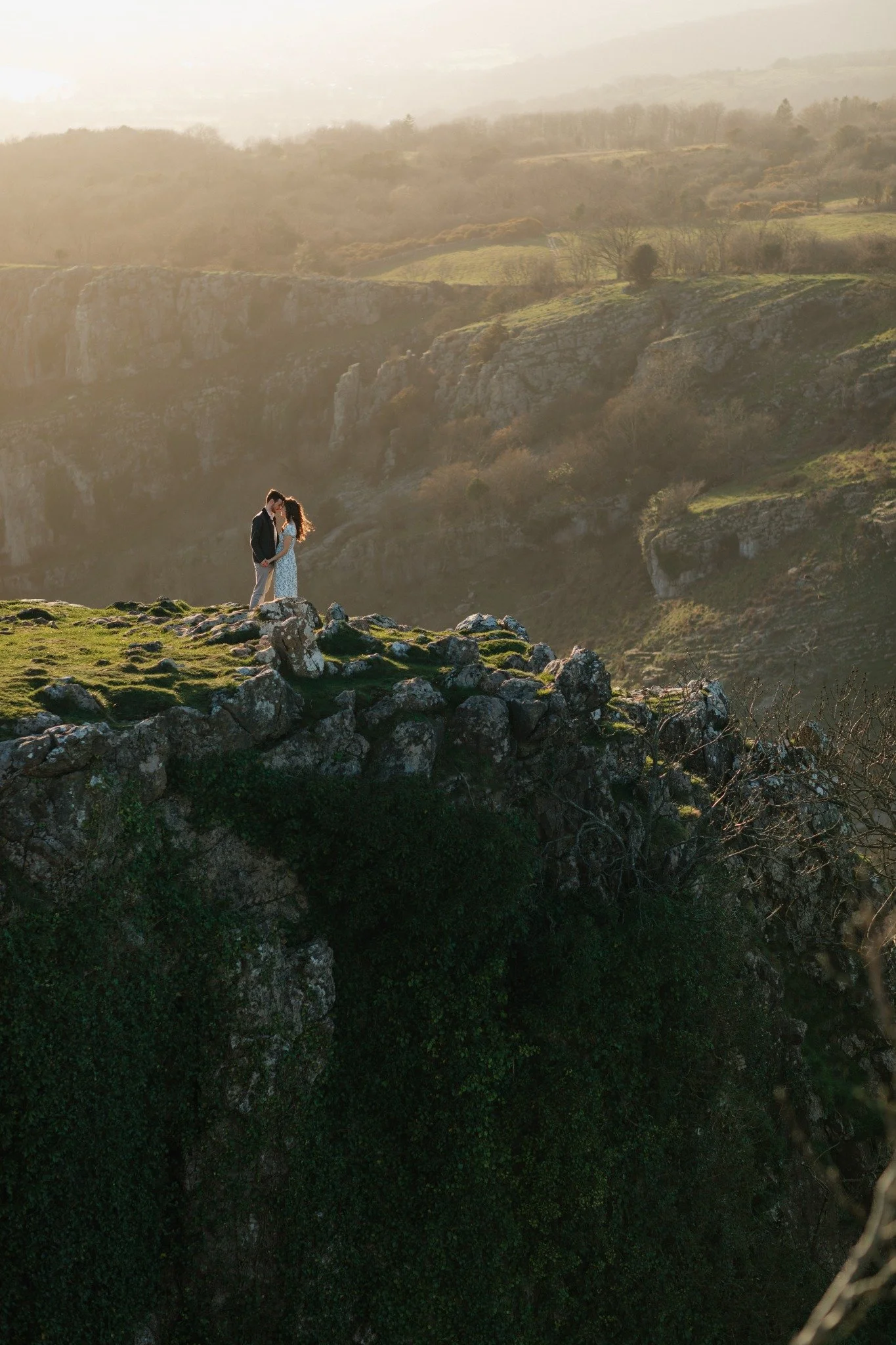 A couple standing on a rocky ledge overlooking a valley with layers of green hills and cliffs, bathed in warm golden sunlight.