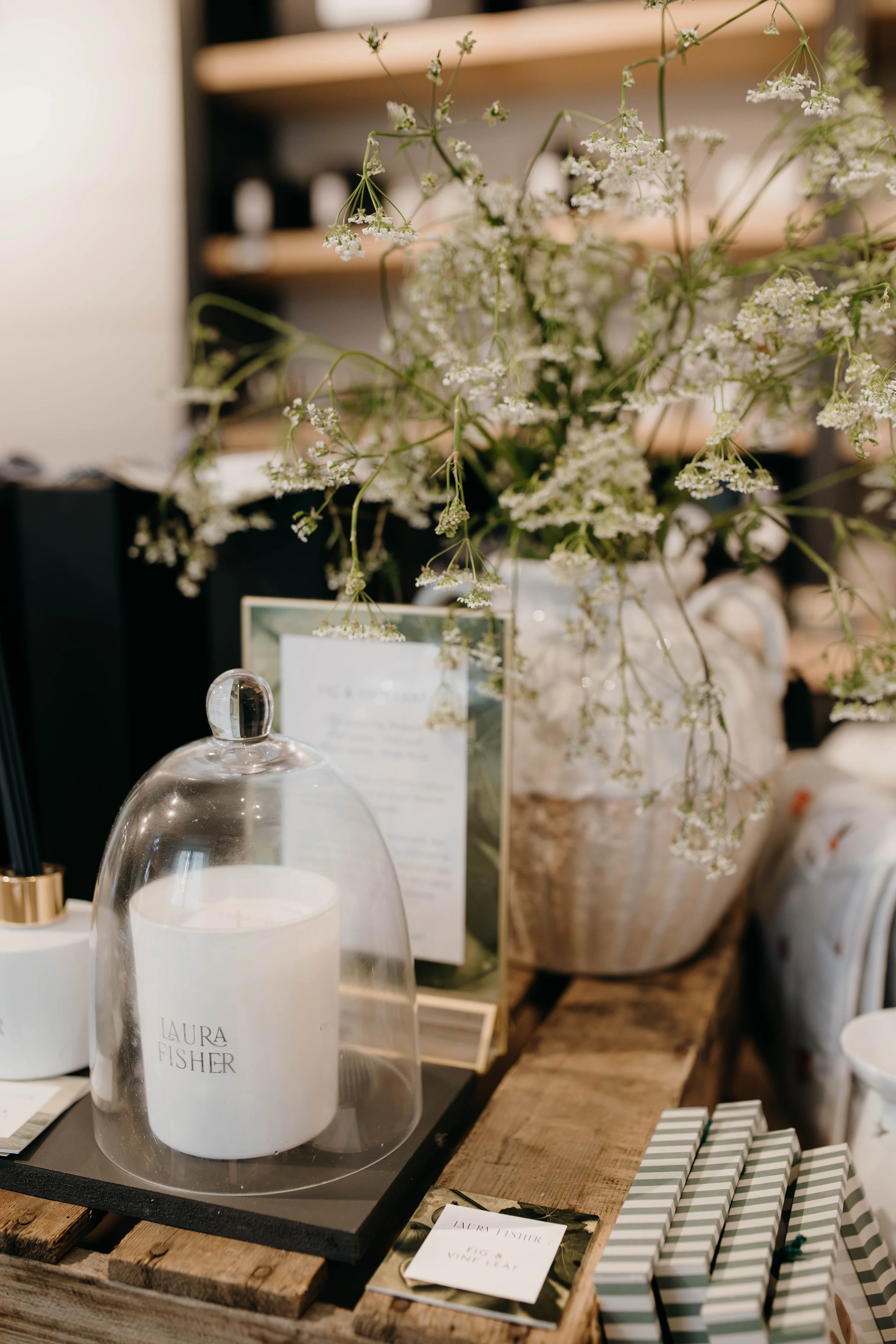 Decorative display featuring a glass cloche with a candle inside, a bouquet of white flowers, and a framed information card on a rustic wooden table in a retail setting.