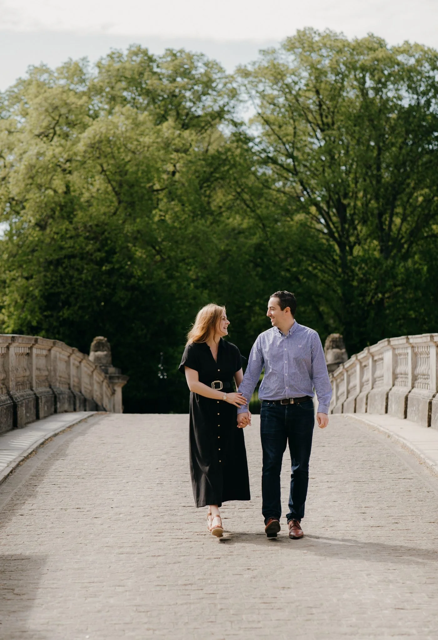 A couple holding hands and walking on a bridge in a park with green trees in the background.