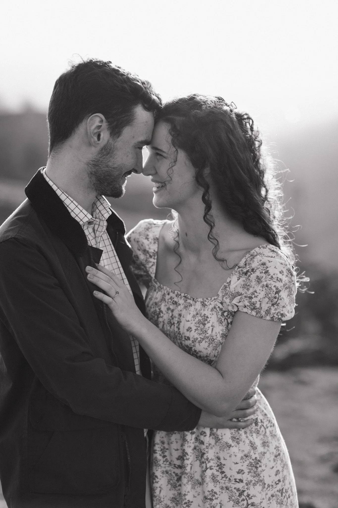 A black and white photo of a couple with foreheads touching, smiling, embracing outdoors with a blurred background.