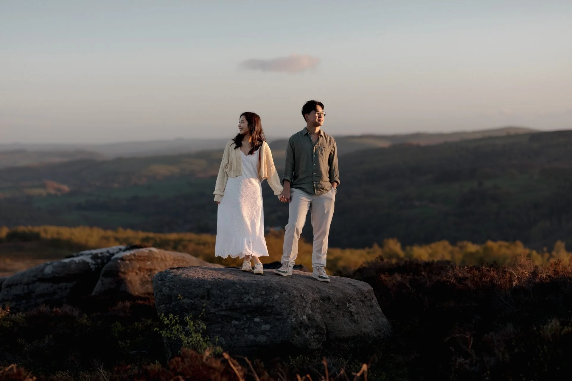A couple holding hands standing on a large rock in a scenic outdoor landscape during sunset, with rolling hills and a cloudy sky in the background.