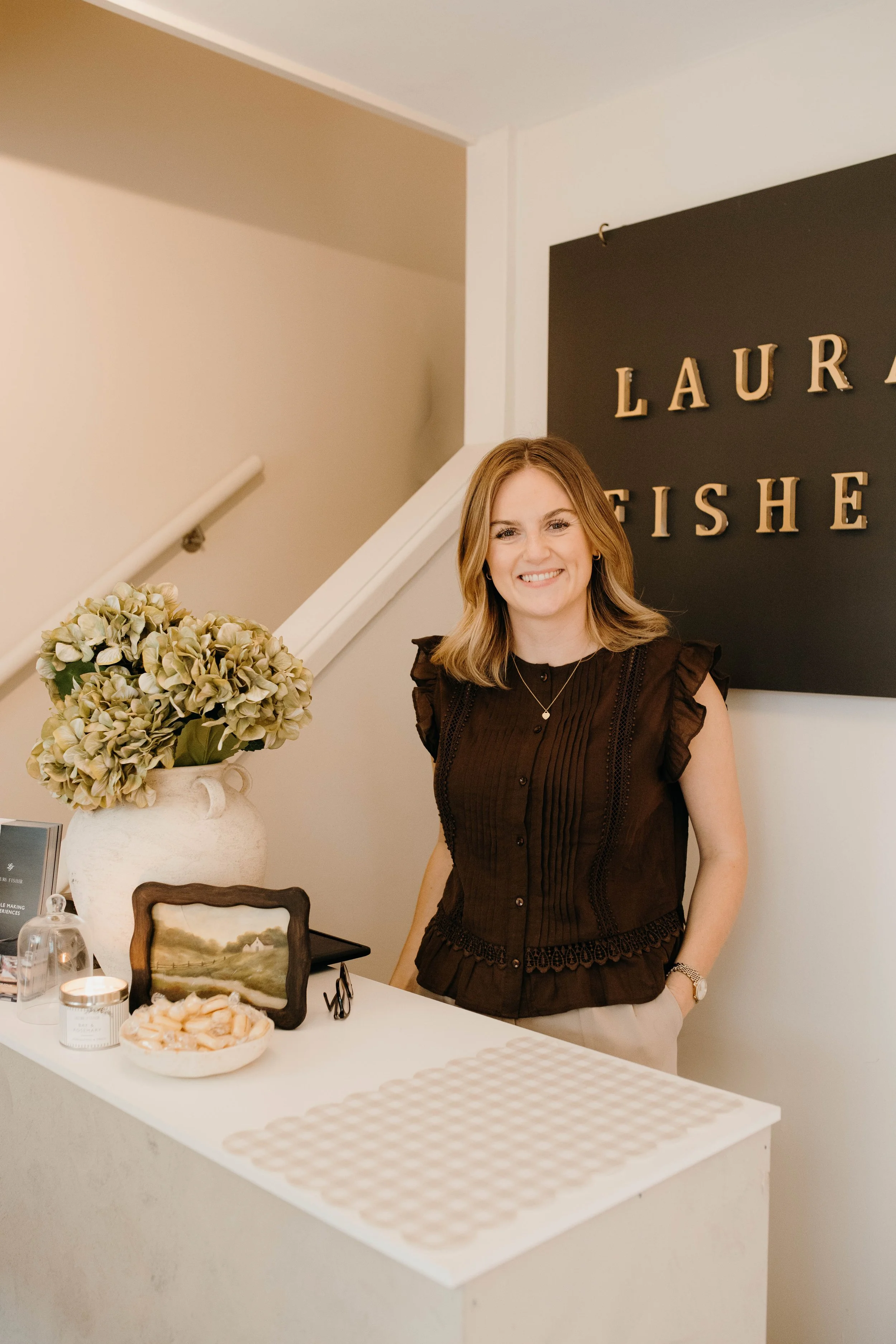 A woman standing behind a white table with decorative items, including a vase with dried hydrangeas, a framed picture, a jar candle, and a bowl of cookies. She is smiling and wearing a brown sleeveless blouse, in front of a black wall with golden let