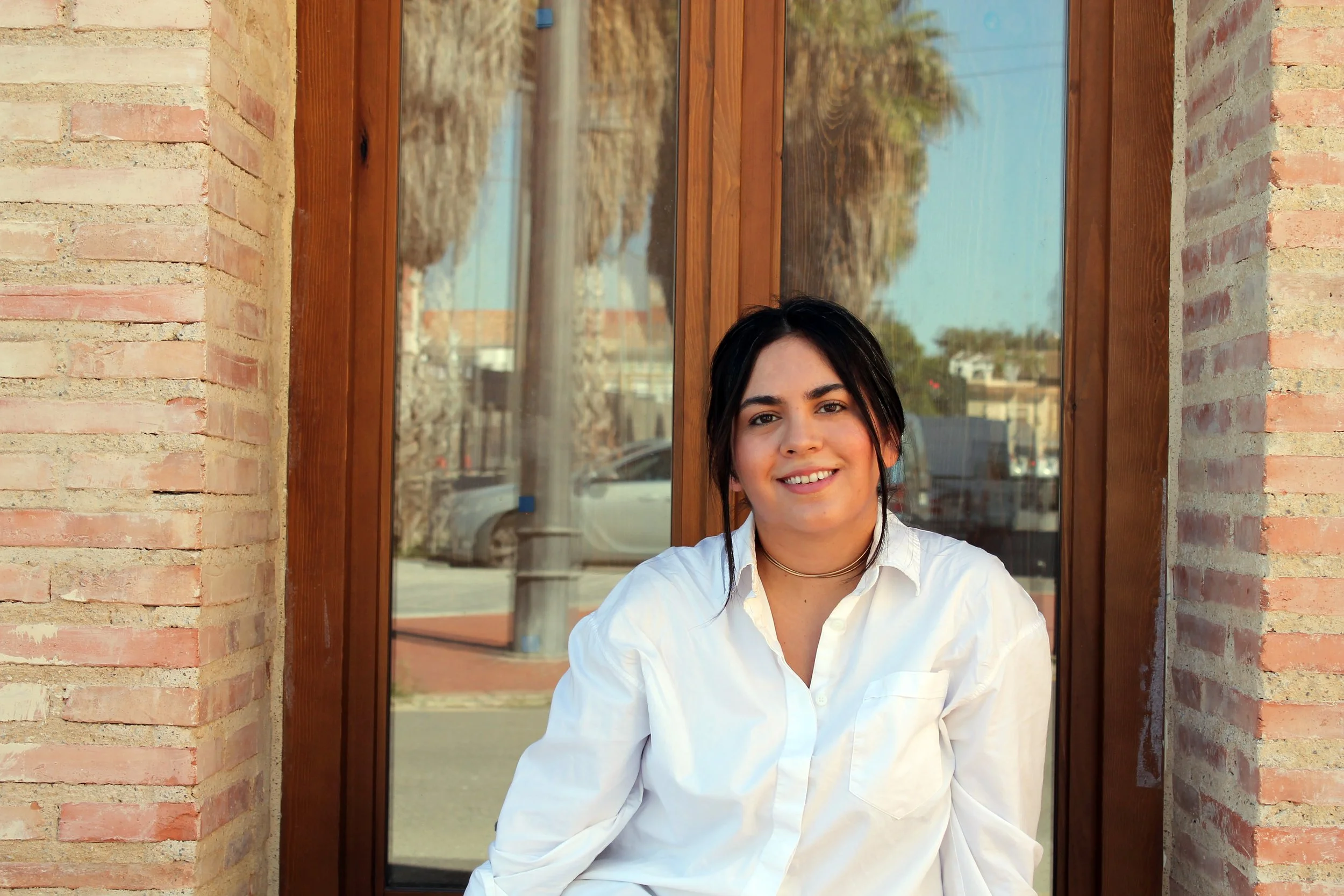 A young woman with dark hair and light skin smiling, sitting outside in front of a glass window with reflections of trees and parked cars.