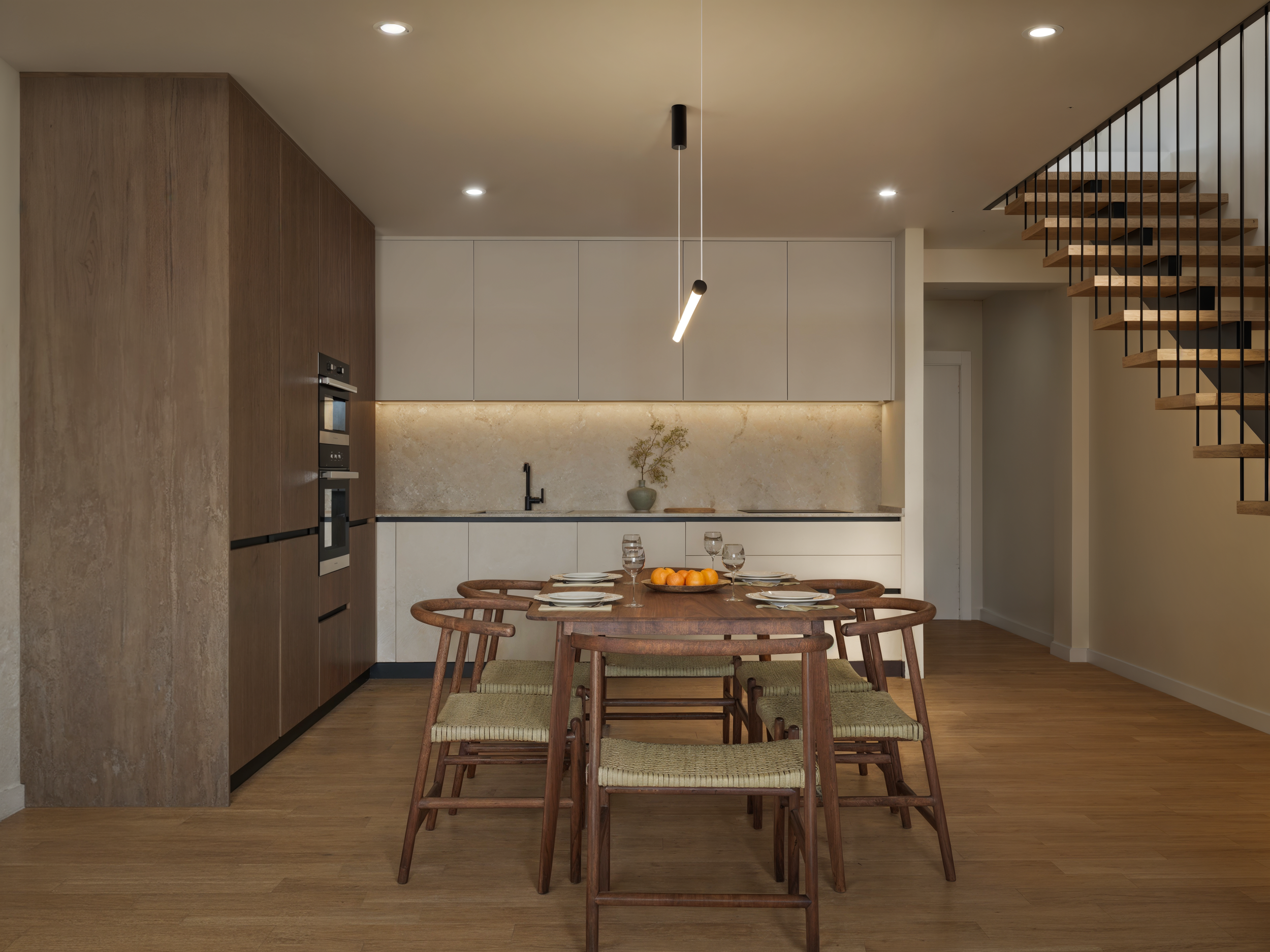 Modern dining area with wooden table and six chairs, set for four with plates, glasses, and a bowl of oranges. Behind it is a sleek kitchen with beige and wood cabinetry, a marble backsplash, and a black faucet. To the right is a staircase with woode