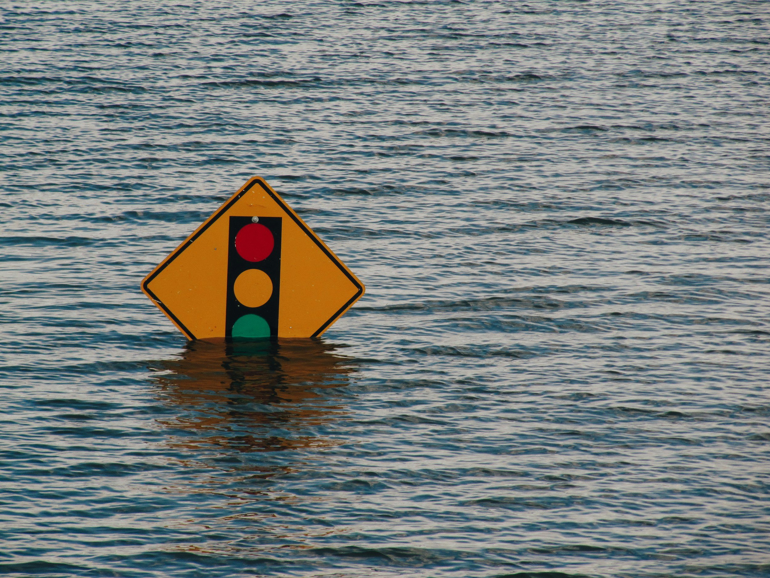 A yellow warning sign with a traffic light symbol partly submerged in water, surrounded by rippling water.