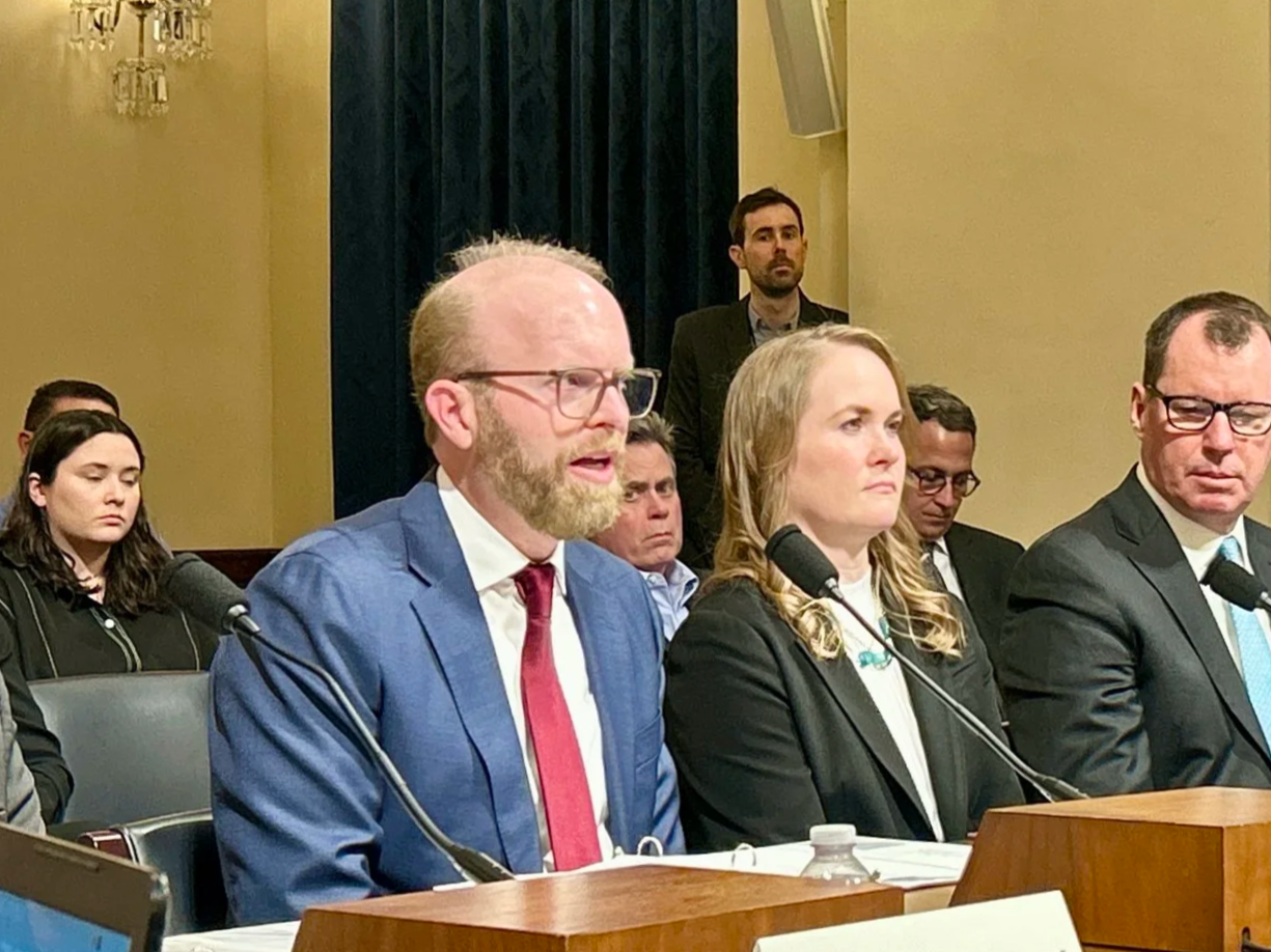 A panel of three people, a man in a blue suit with glasses and a beard speaking, a woman with blonde hair, and a man with glasses, sit at a table with microphones during a formal meeting or hearing. Several other people are seated behind them in the background.