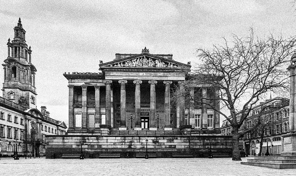 A historic neoclassical building with tall columns, intricate pediment sculpture, and a staircase in front, surrounded by other classic architecture, leafless trees, and an overcast sky.