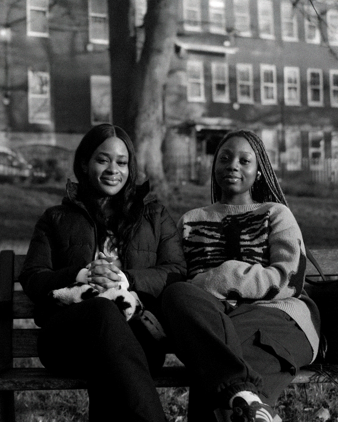 Two young women sitting on a park bench with a background of a tree and buildings, smiling at the camera in a black and white photograph.