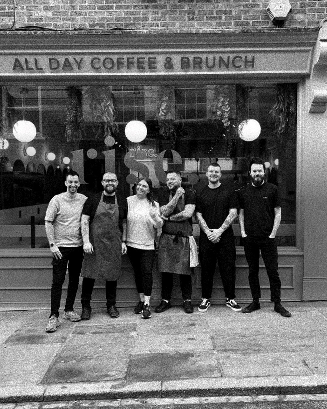 Group of six people standing in front of a restaurant window, some wearing aprons, with a sign above that reads 'All Day Coffee & Brunch'.