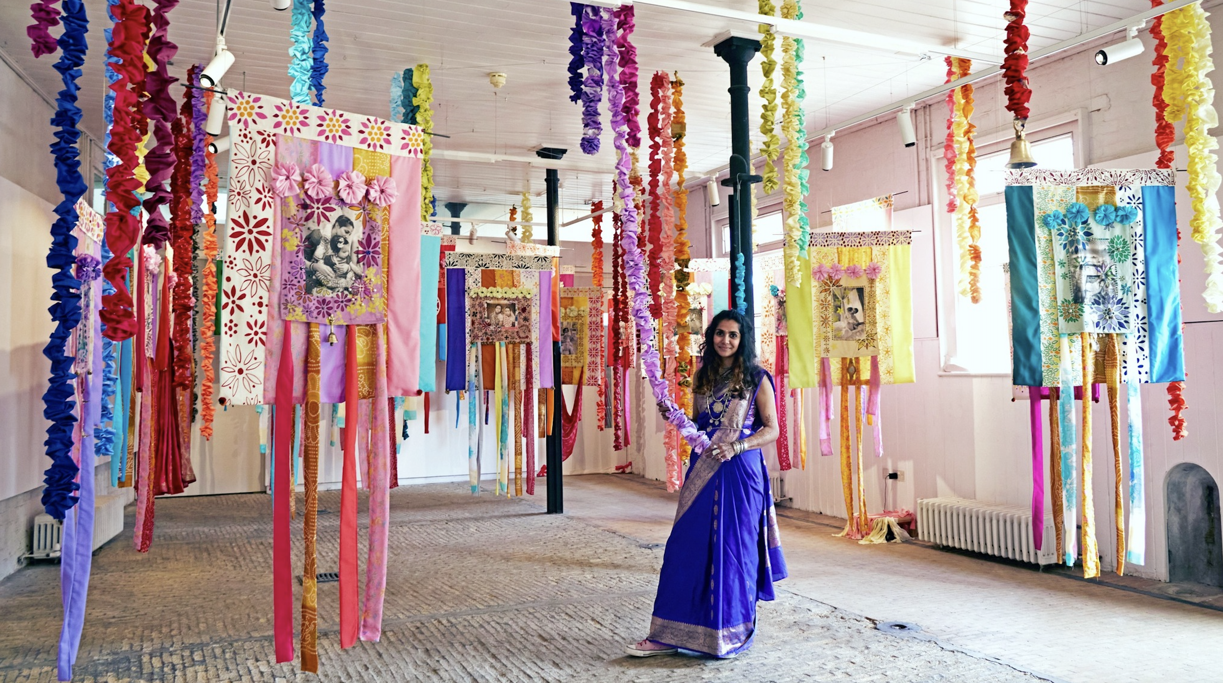 A woman in a purple saree standing in a room decorated with colorful hanging fabric and artwork for a celebration or cultural event.