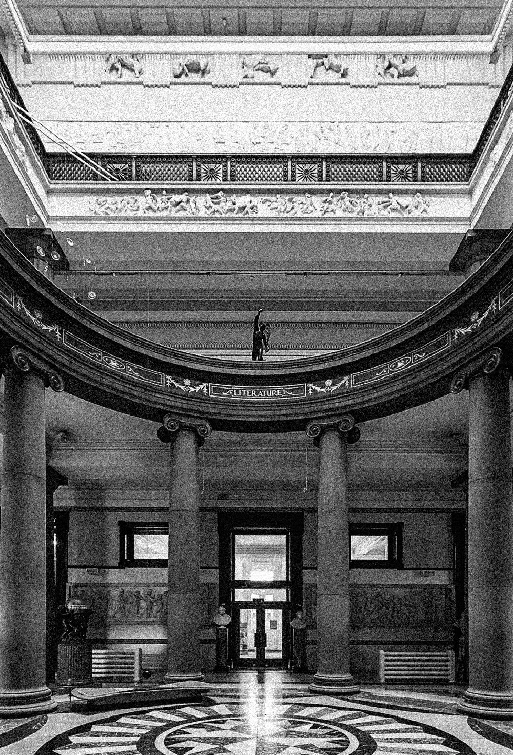 Interior of a grand building with columns, a circular patterned floor, and multiple floors with decorative railings and sculptural details. A person is seen on the upper level, and two security personnel stand at the entrance.