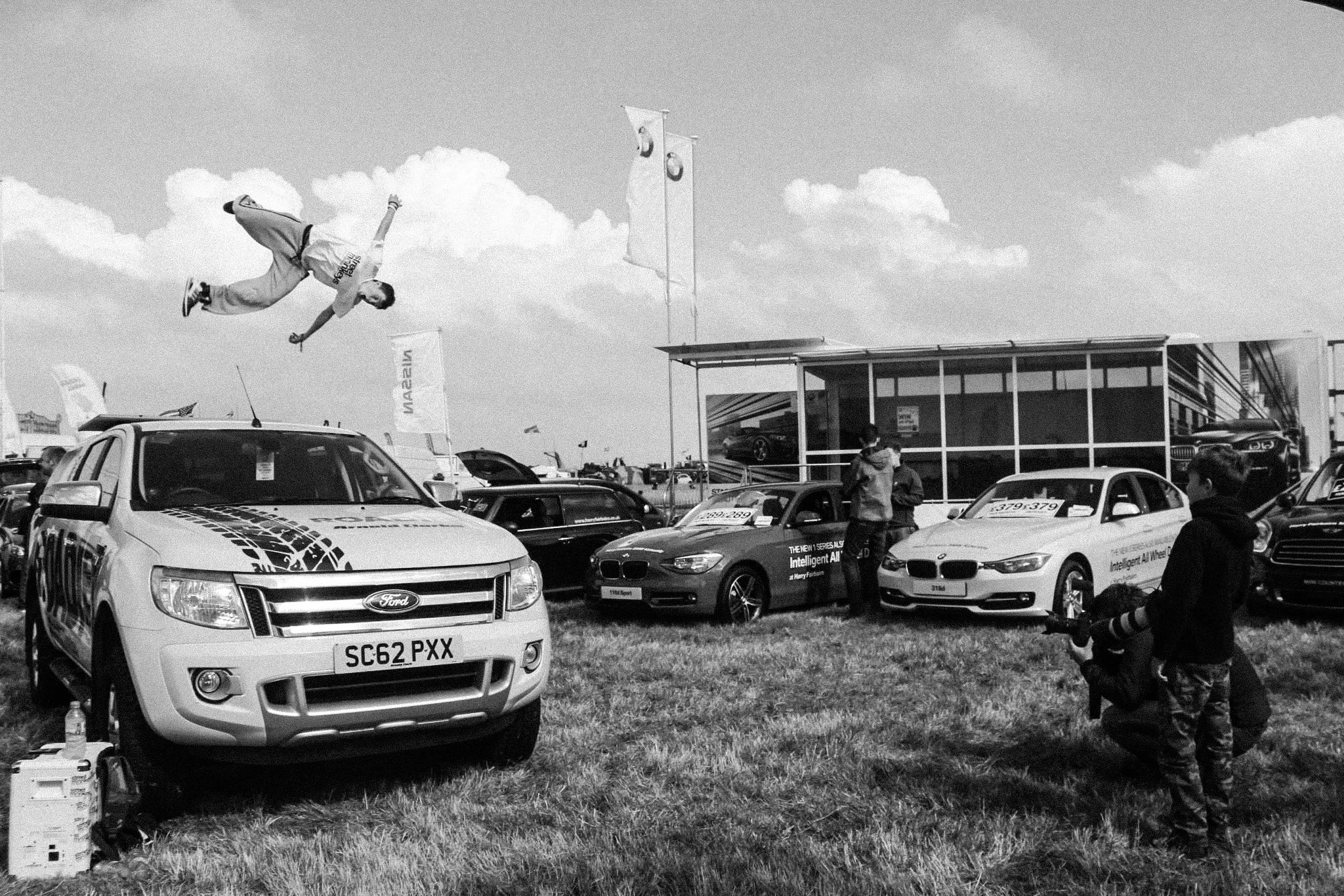 A person is mid-air performing a stunt on top of a Ford vehicle at an outdoor event, with several other cars and people around.