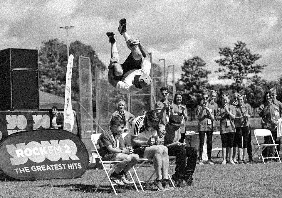 A person performing a backflip in mid-air at an outdoor event, with a group of spectators watching.