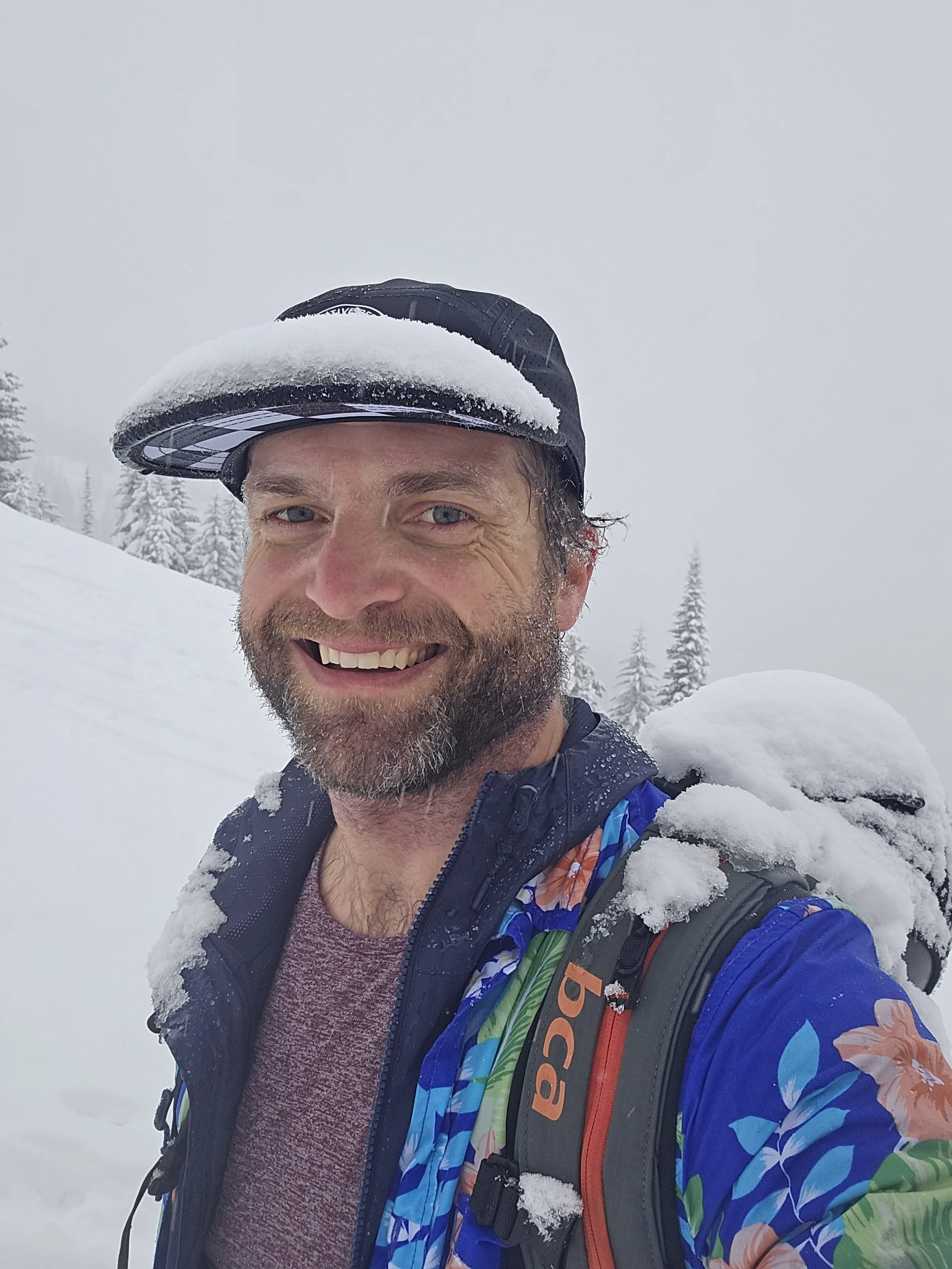 A smiling man with a beard wearing a black cap with snow on it, a colorful floral jacket, and a backpack, standing in a snowy landscape with snow-covered trees in the background participating in a fundraiser for DREAM in Whitefish, MT.