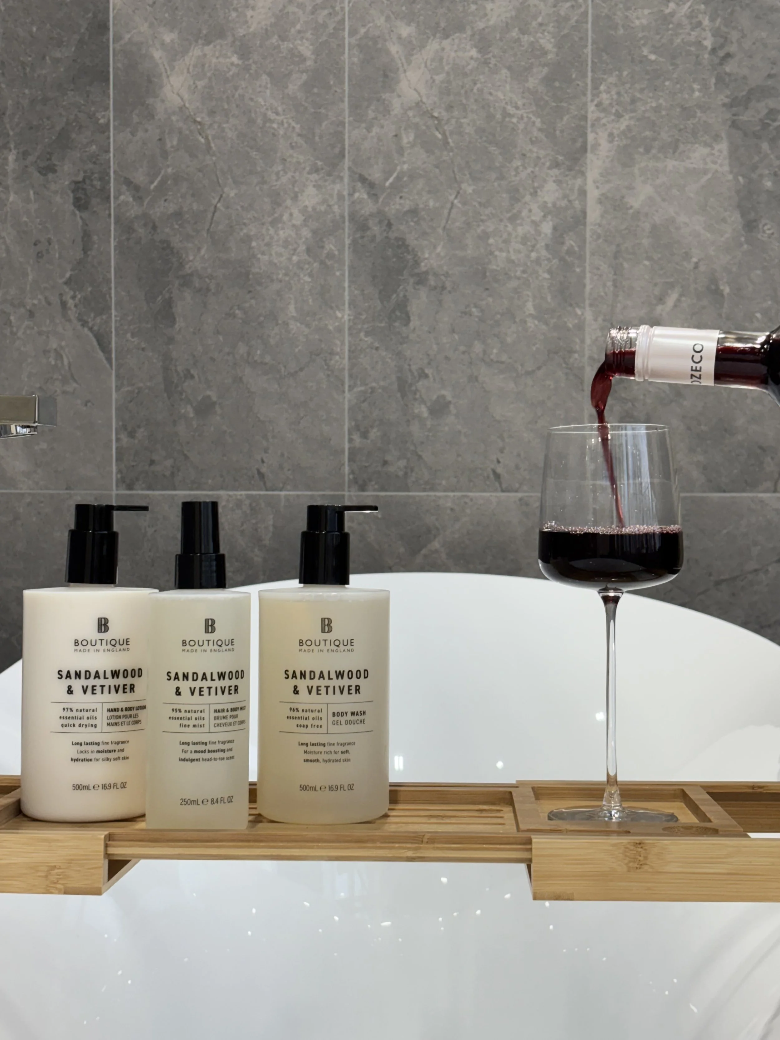 Three bottles of Sandalwood & Vetiver skincare products and a glass of red wine being poured from a bottle, all arranged on a wooden tray over a white bathtub with a gray tiled wall in the background.