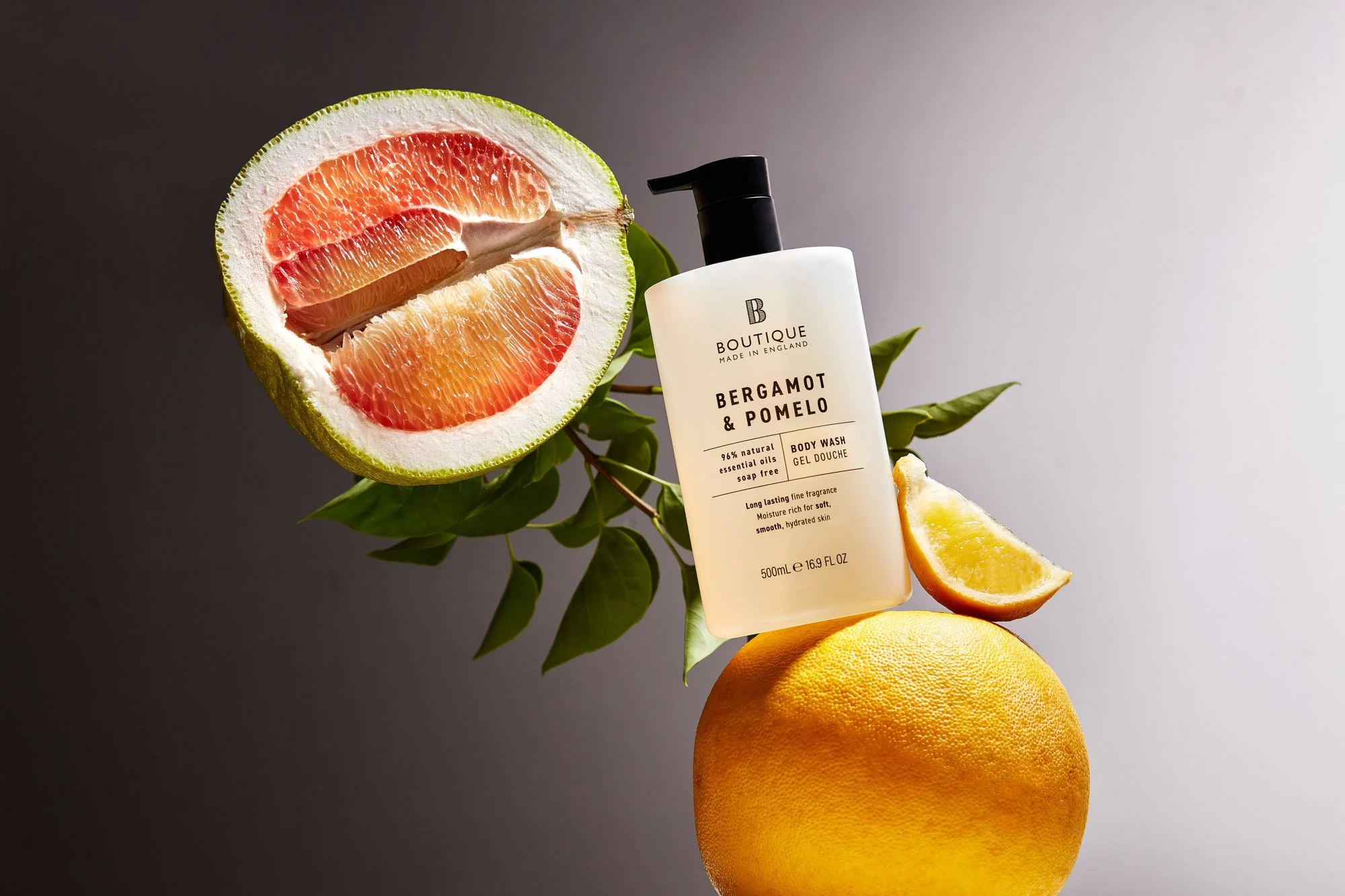 Bottle of body wash labeled Bergamot & Pomelo surrounded by grapefruit, lemon, and grapefruit half, with green leaves on a neutral background.