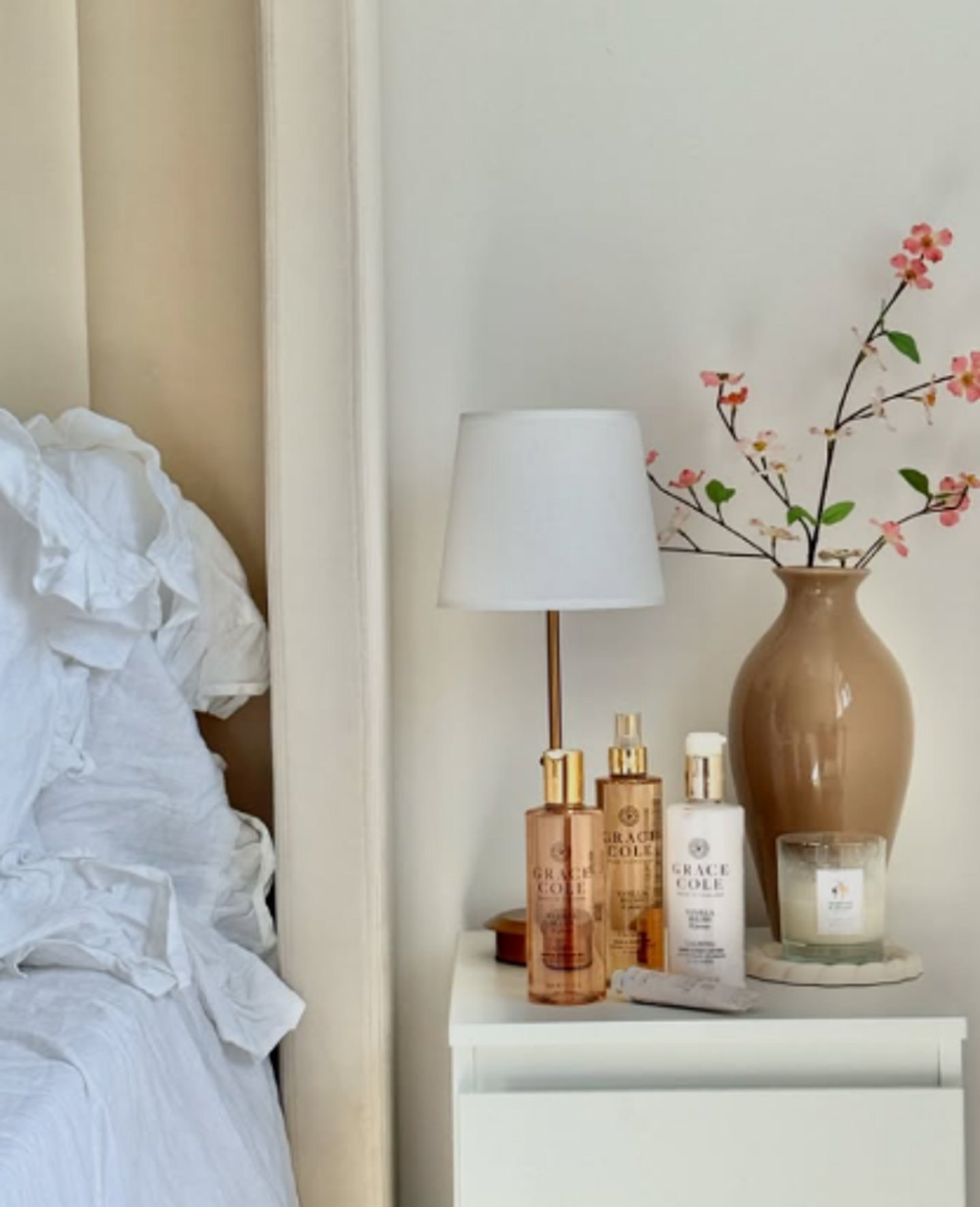 Bedroom nightstand with a lamp, a large brown vase with pink artificial flowers, Grace Cole skincare bottles, a lit candle, and a small white rolled towel.