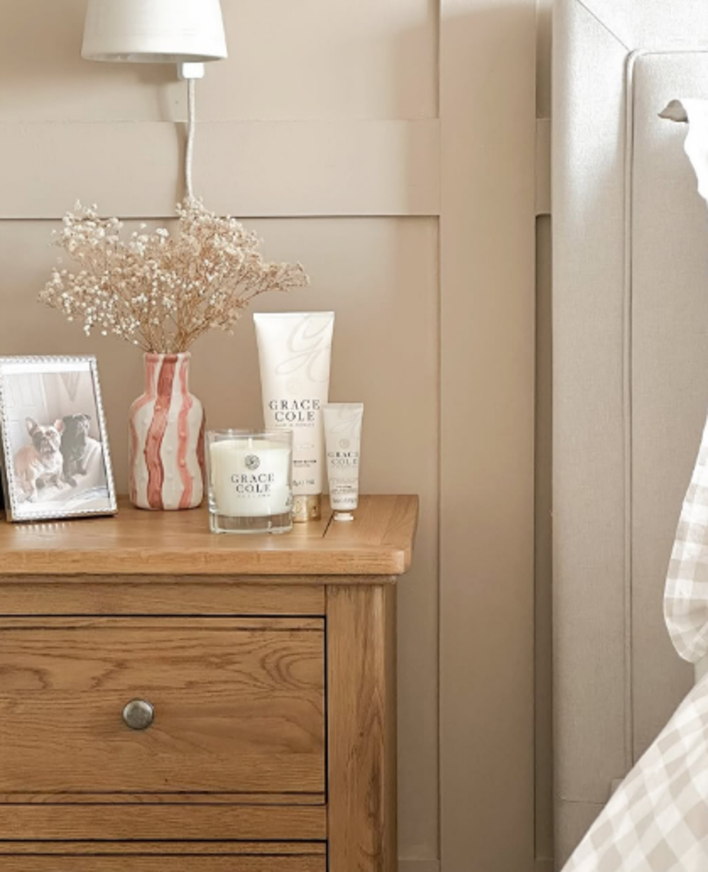 A wooden nightstand with a framed photo of two dogs, a pink and white vase with dried flowers, a Grace Cole scented candle, and skincare products, located next to a beige bed and wall-mounted lamp.
