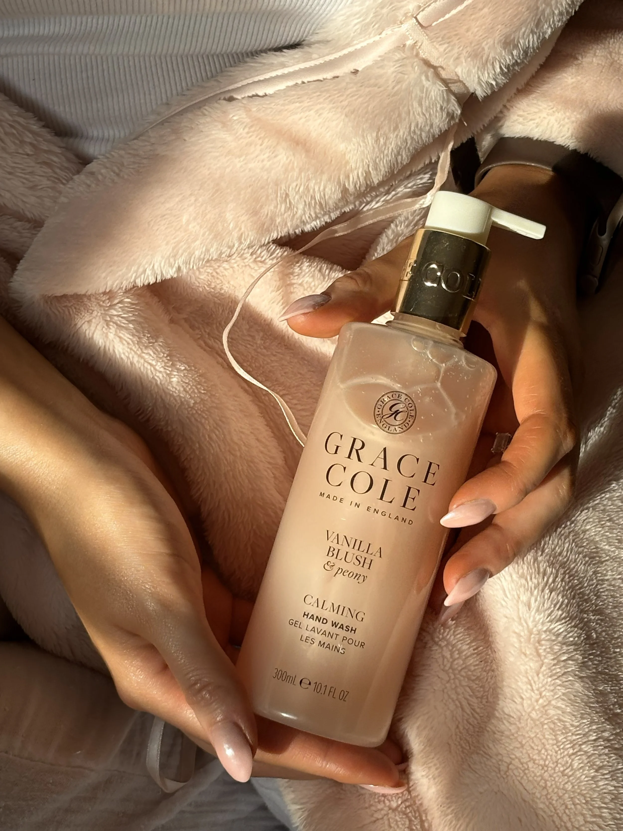 A person holding a bottle of Grace Cole Vanilla Blush & Peony calming hand wash, wearing a beige fleece jacket and sitting on a beige surface.