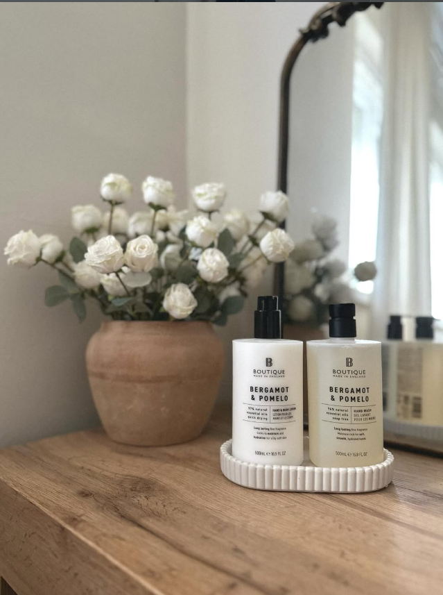 A wooden dresser with a potted white flower arrangement and two bottles of Bergamot & Pomelo hand and body products on a white tray, with a mirror and a window with curtains in the background.