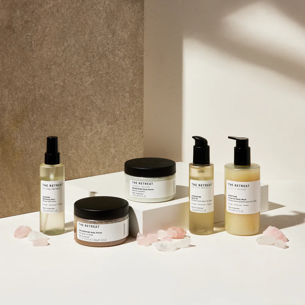 Beauty skincare products including serums, body lotion, body polish, and a jar of cream, arranged with pink and white crystals on a white surface with shadows.