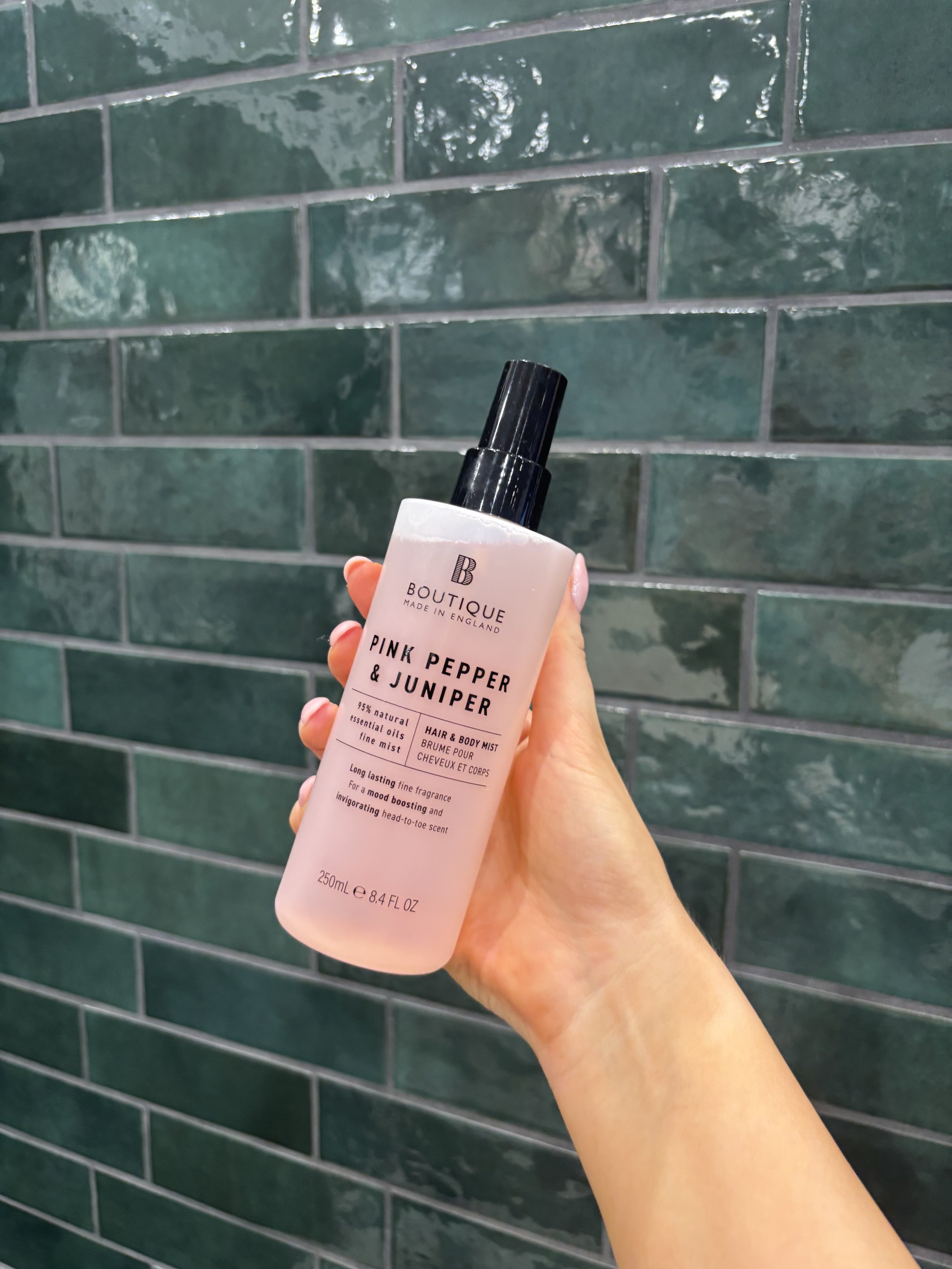 A person holding a pink bottle of 'Pink Pepper & Juniper' hair and body mist against a green tiled wall.