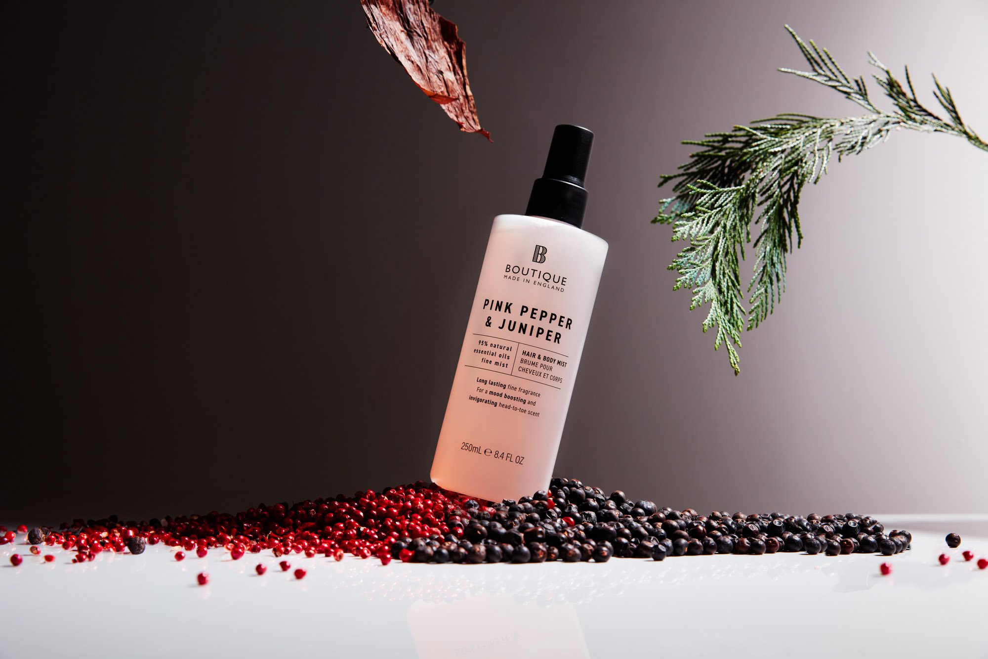 A bottle of Boutique Pink Pepper & Juniper hair and body mist leaning against black and red berries on a white surface with green pine branches and a partial red leaf as background.