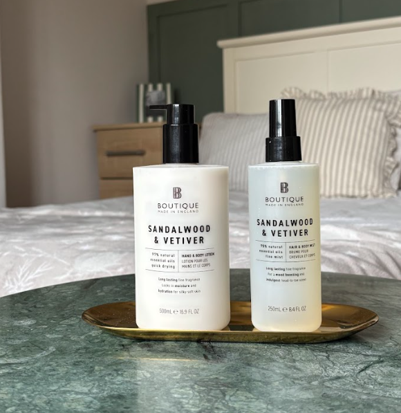 Two bottles of sandalwood and vetiver scented lotion on a gold tray in front of a bed with striped pillows in a bedroom.