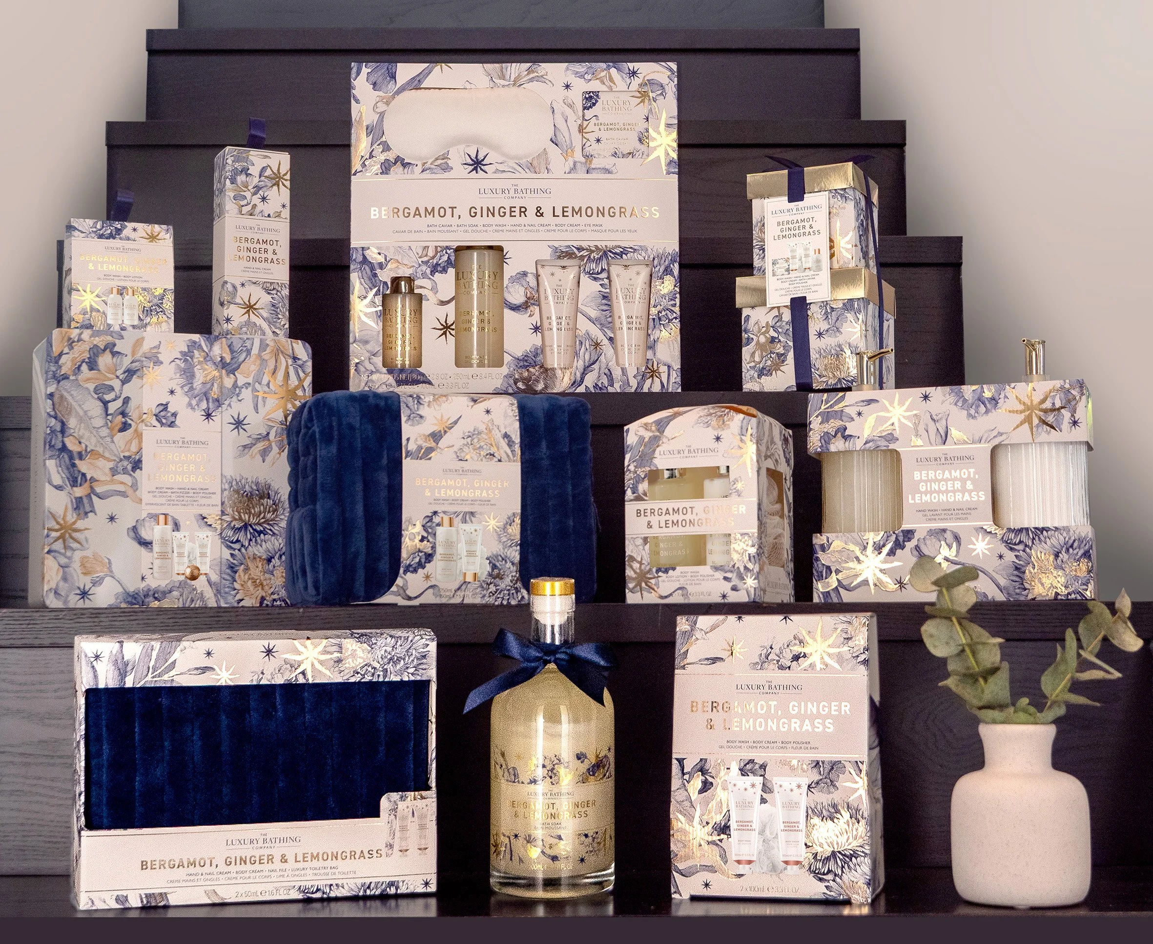 Display of luxury bath products with bergamot, ginger, and lemongrass scent, decorated with floral and starry patterns, arranged on dark shelves with a small potted eucalyptus plant in a white vase.