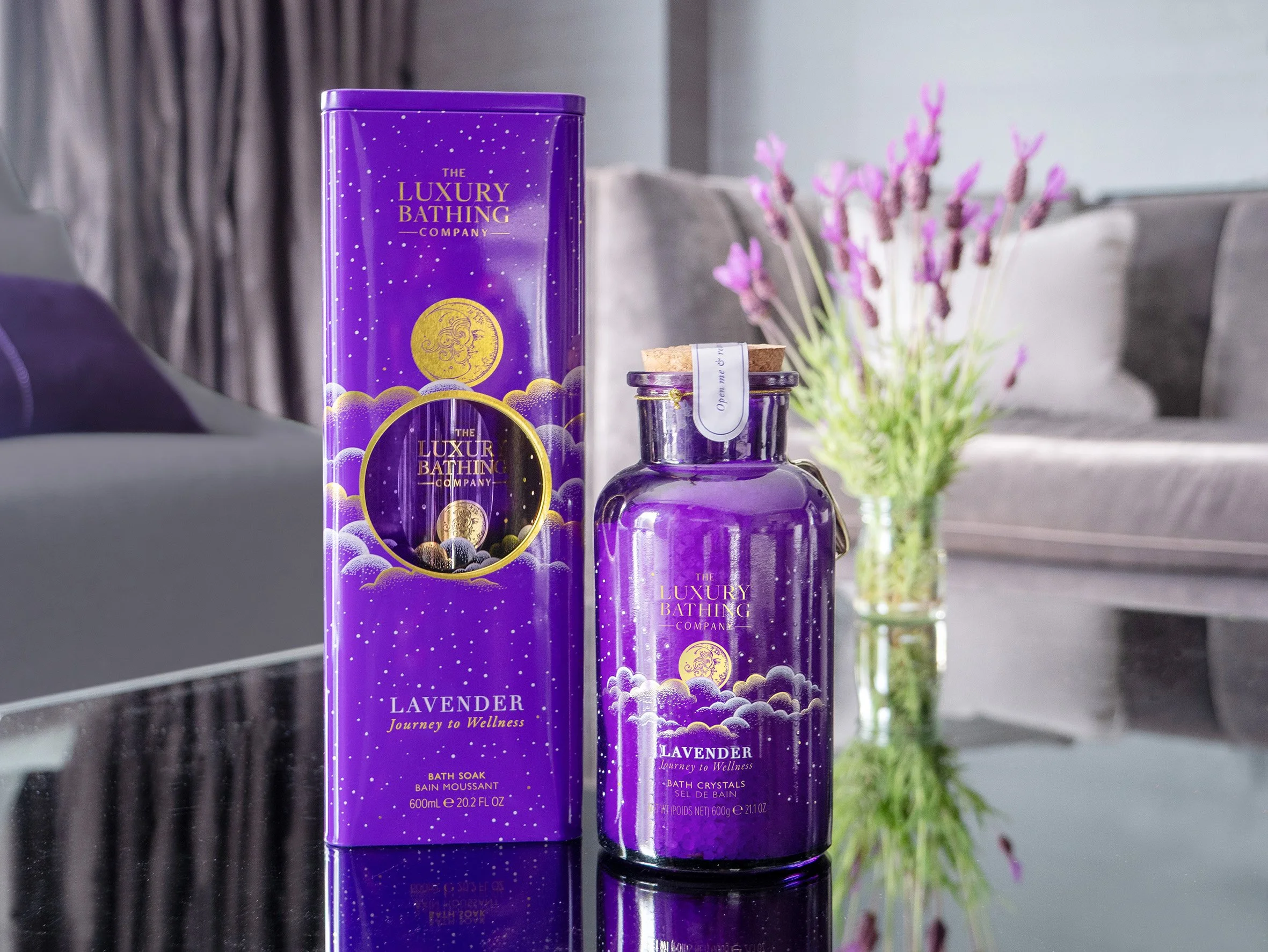 A purple box and a purple bottle of lavender bath soak from The Luxury Bathing Company on a reflective black surface, with a glass vase of pink flowers in the background.