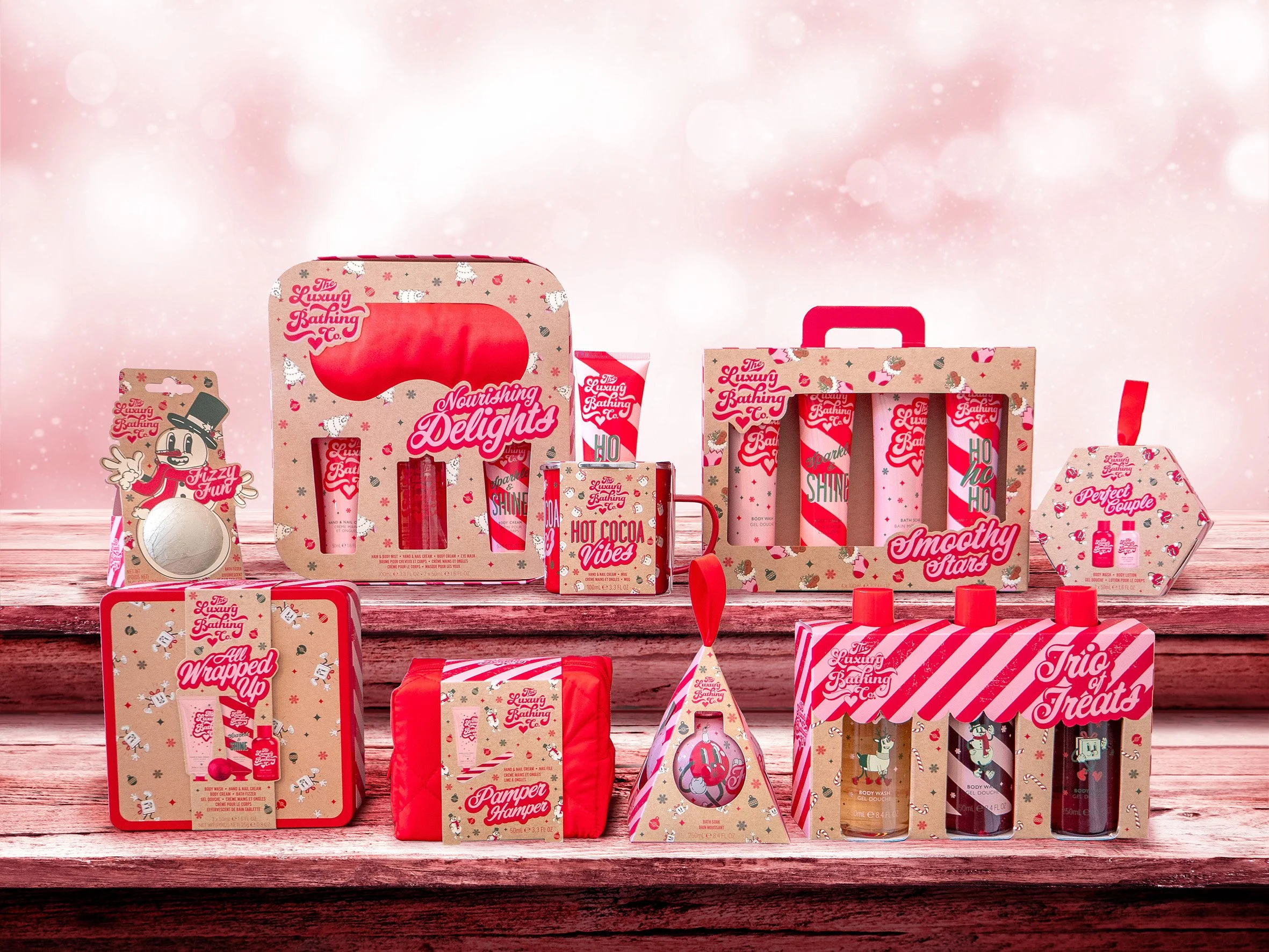 Collection of holiday-themed bath and body products with red, pink, and white packaging, displayed on a wooden surface against a pink bokeh background.