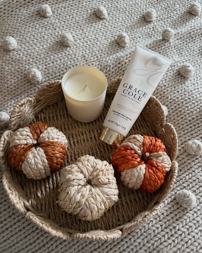 A candle, a tube of Grace Cole body butter, and four small decorative pumpkins on a woven tray, all on a chunky knit blanket with pom-poms.