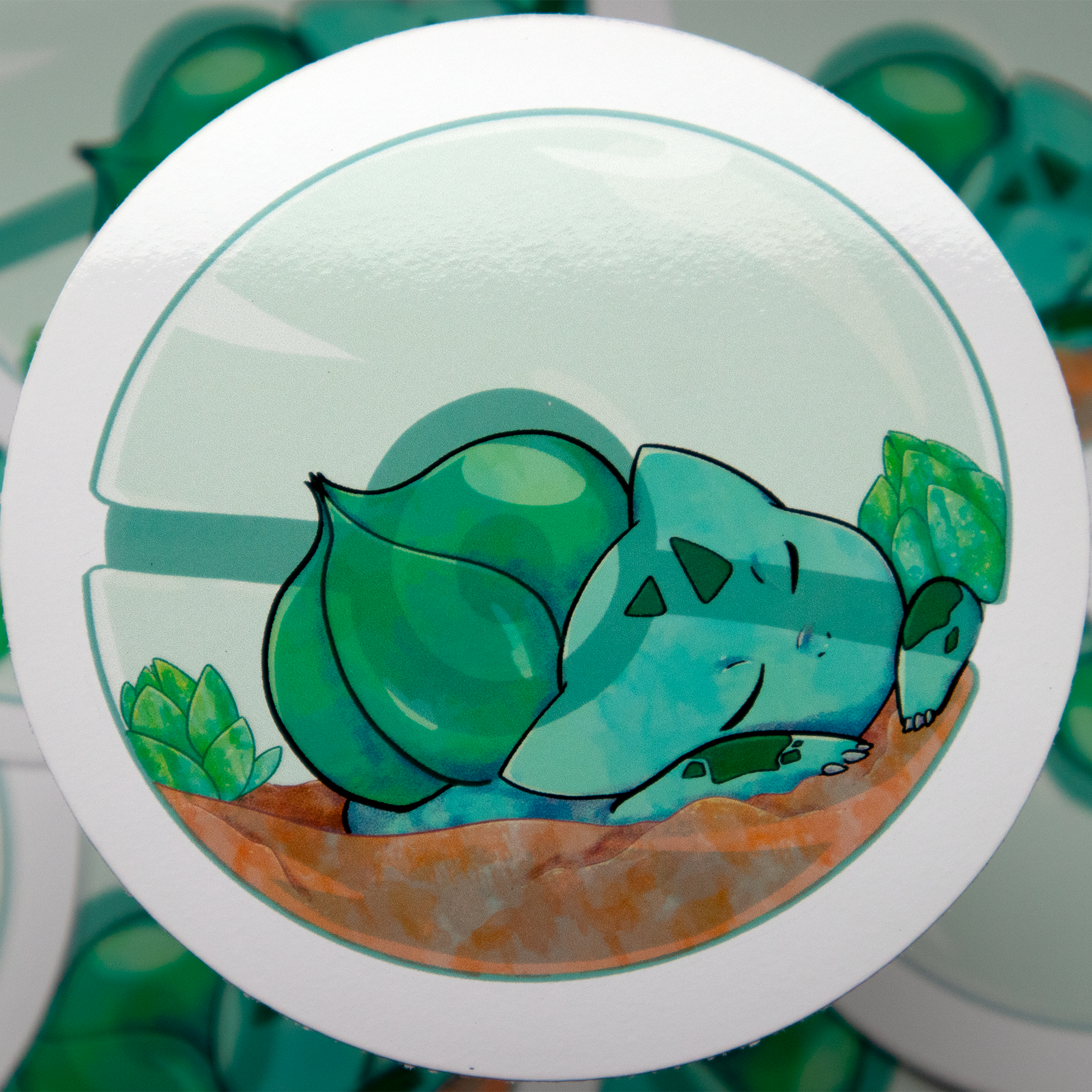 A cartoon illustration of 'Bulbasaur' from Pokémon, a green dinosaur with a flower on its back. It's eyes are closed, resting on soil, surrounded by green plants in a Pokeball.