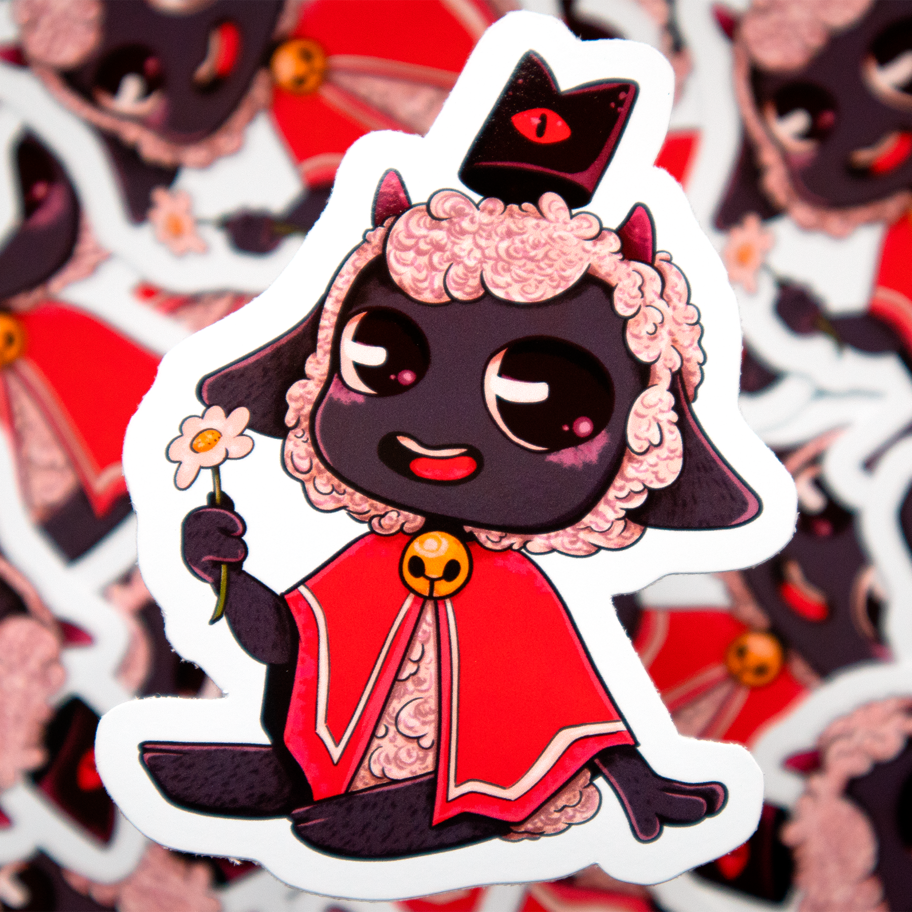 Cute cartoon lamb from 'Çult of the Lamb' video game. It's got a dark face with white wool, two little horns, a black hat and a red cape with a yellow bell around it's neck. It's sitting and holding a flower with a smile.