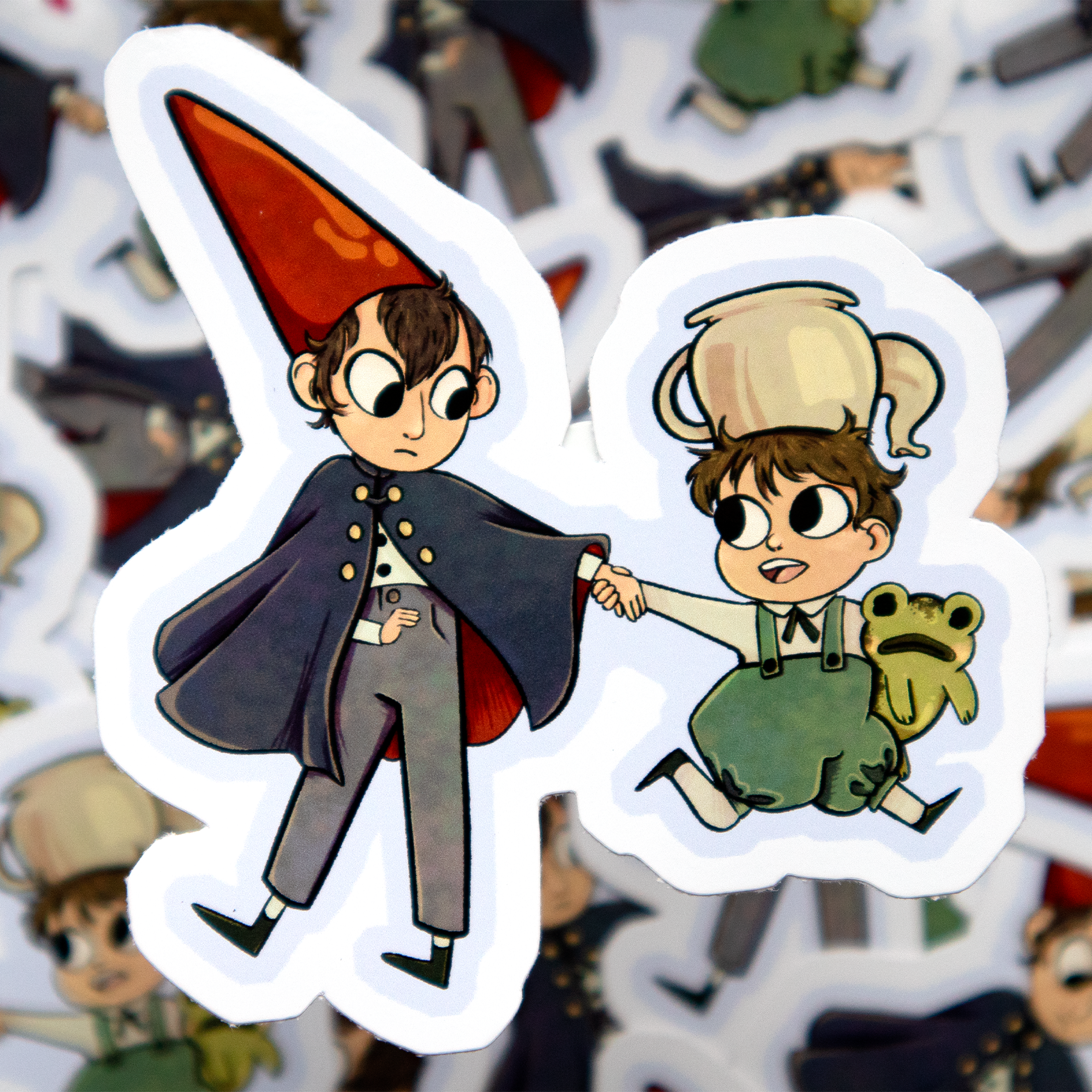 Sticker of two cartoon boys shaking hands, one dressed as a gnome with a tall red hat and cape, the other wearing a tea pot hat and holding a small green frog. Greg and Wirt from the Cartoon network Series 'Over the Garden Wall'