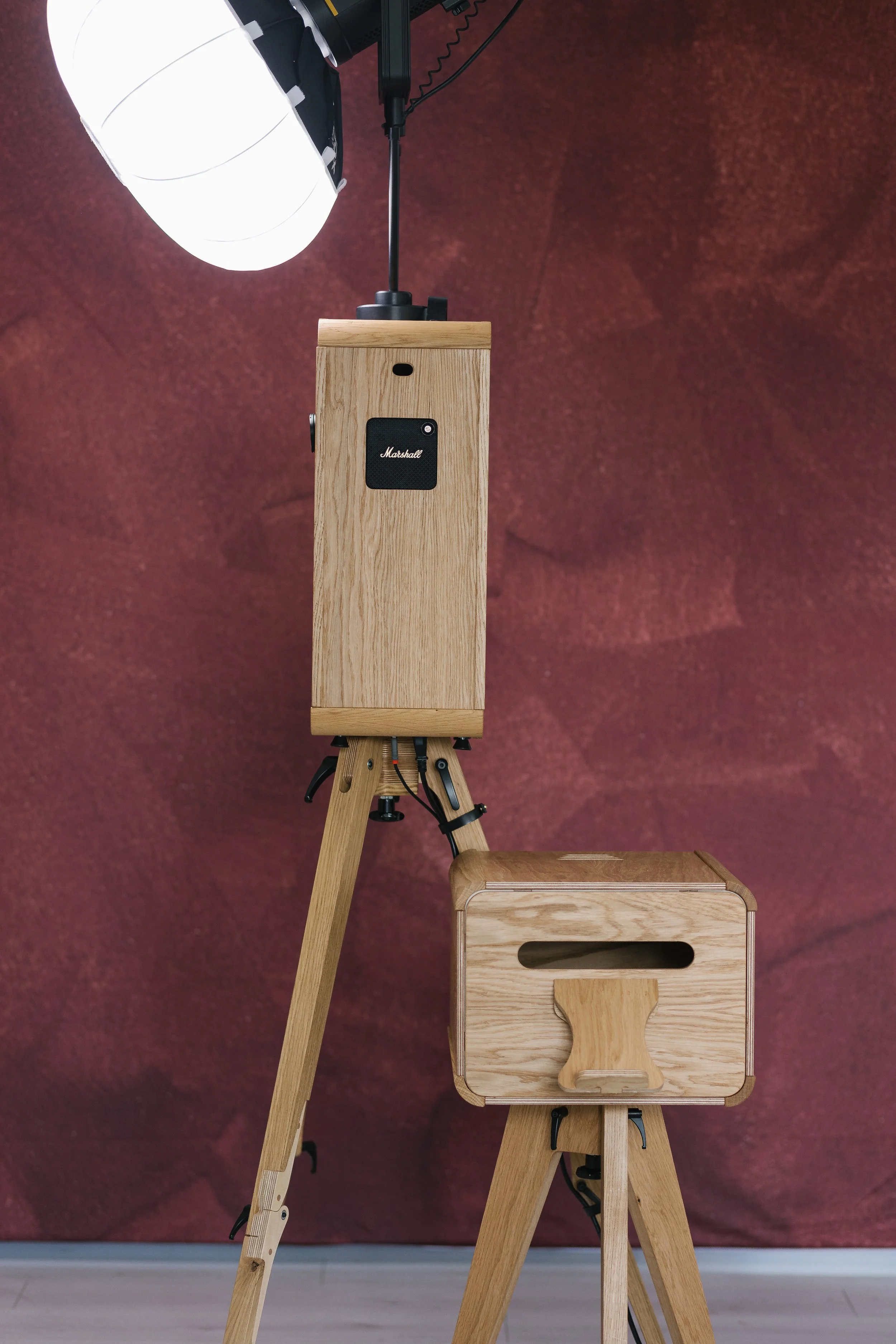 A wooden speaker mounted on a tripod stand and a wooden box with a handle, also on a tripod stand, with a studio light hanging above against a reddish-brown backdrop.