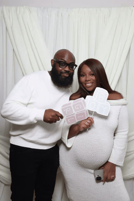 A couple celebrating a pregnancy, holding signs that say "Baby Shower" in front of a white curtain. The woman is pregnant, and the man is wearing glasses and a white sweater.