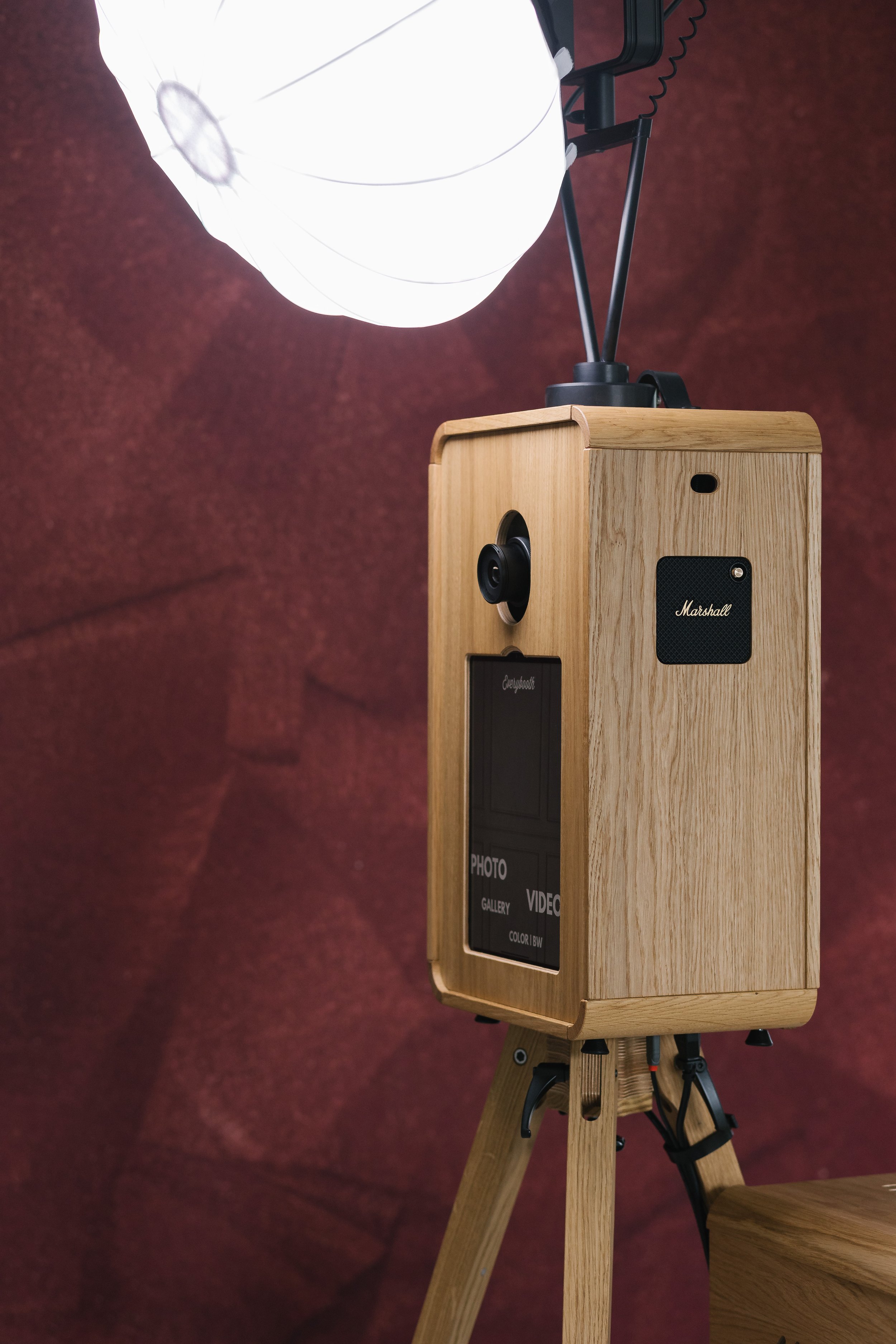 A vintage-style photo booth with a wooden casing, mounted on a tripod, equipped with a camera lens, a display screen, and a speaker, positioned in front of a maroon textured background, illuminated by a large white lamp