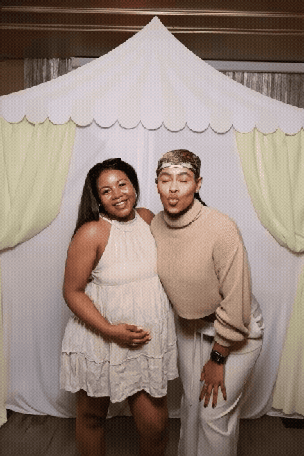 Two women posing in front of a decorative backdrop resembling a small house with a pointed roof and curtains, one smiling and one making a playful face.