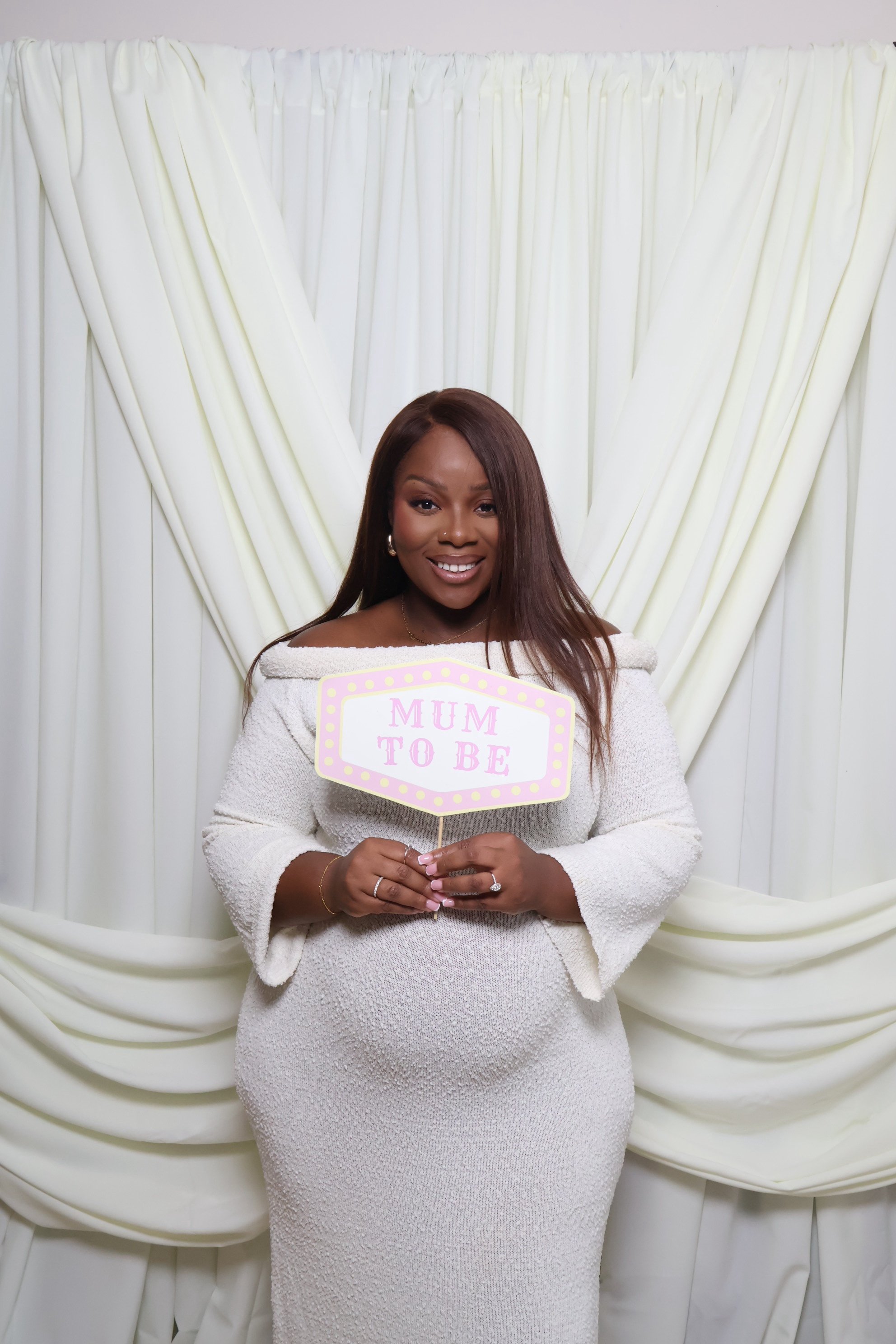 Pregnant woman smiling and holding a sign that says 'MUM TO BE' in front of a cream-colored curtain backdrop.